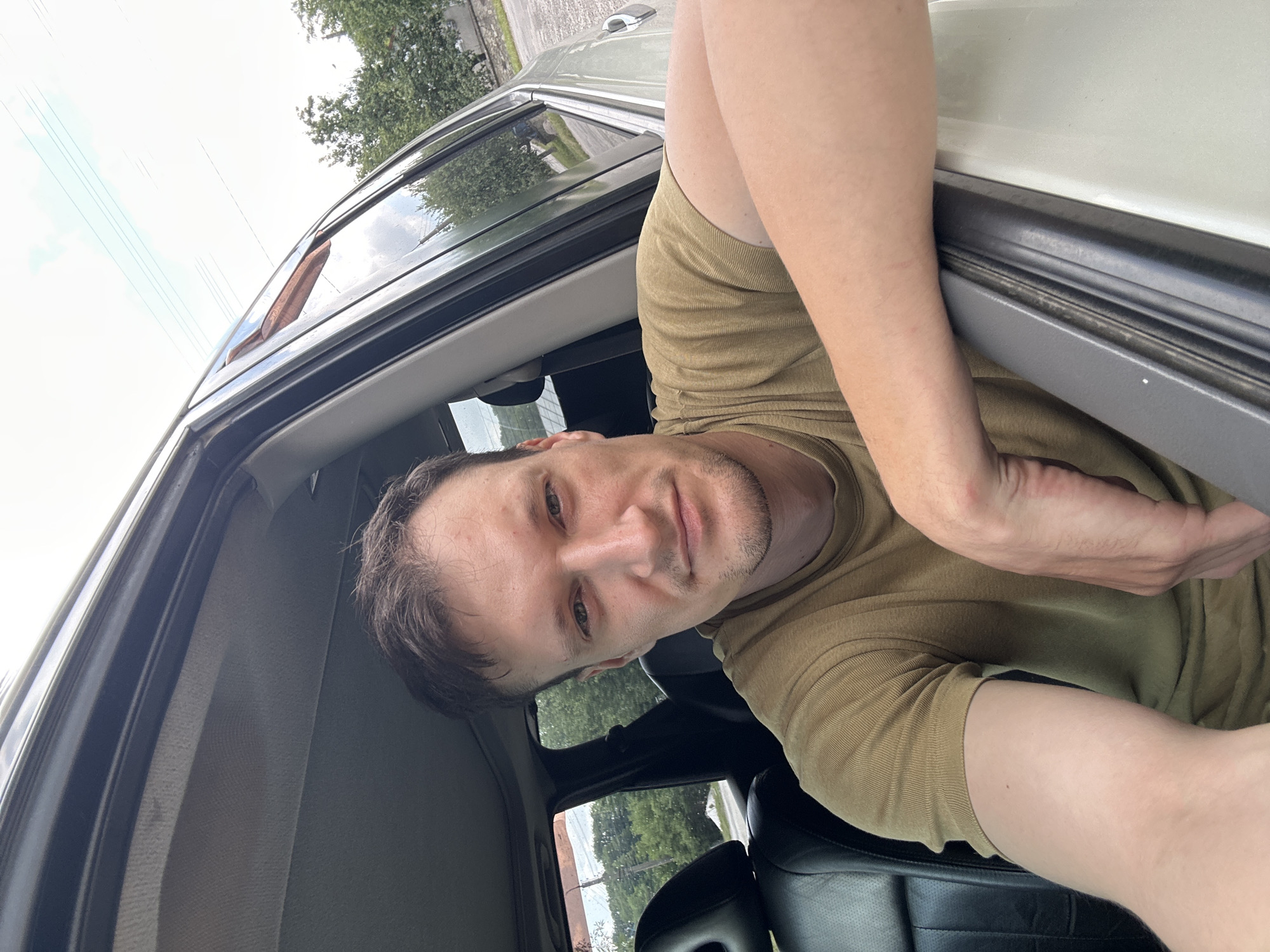 A man in a tan t-shirt is sitting in the driver's seat of a light-colored SUV, looking at the camera. He has short brown hair and a light beard. His left arm is resting on the open car door, and his right arm is bent in front of him. The interior of the car is visible, including black leather seats. Outside the car, trees and a cloudy sky are visible. The scene appears to be in a rural or suburban area, possibly on a road or driveway. The provided location is Makiv, Ukraine, but this is not visually verifiable from the image.