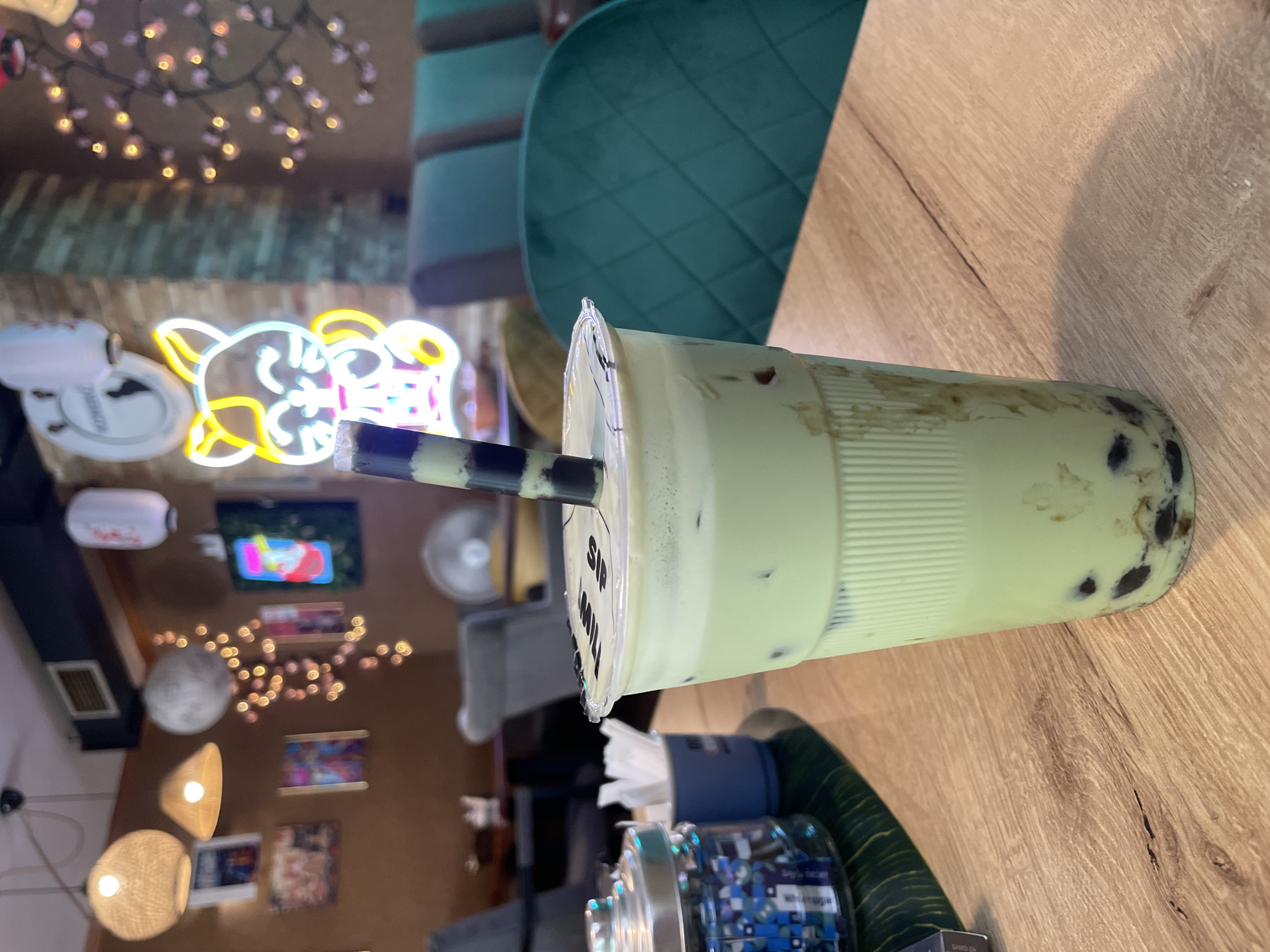 A green-colored beverage with tapioca pearls is displayed in a clear plastic cup with a sealed lid and a striped straw. The cup is placed on a light-colored wooden table. In the background, several decorative elements are visible, including a neon cat sign, string lights, and framed artwork. The setting appears to be an indoor establishment, possibly a cafe or a restaurant, with comfortable seating in teal upholstery visible. The overall ambiance suggests a trendy and inviting space. The specific location in Podgorica, Montenegro, cannot be determined from the visual information alone.