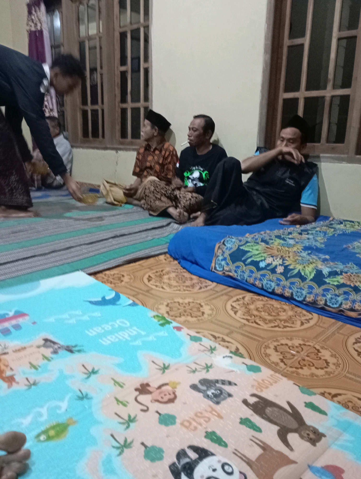 The image depicts an indoor scene, likely a home in Ketepung, Indonesia, with several individuals present. The floor is covered with patterned mats and a blue mat with depictions of continents and animals. In the foreground, a blue mattress with a floral patterned blanket is visible.
Three men are seated on the floor, facing towards the left of the frame. The man on the far right wears a black vest over a blue shirt and a black cap. He appears to be gesturing with his right hand. The middle man is wearing a black t-shirt and has a cigarette in his mouth, looking towards the left. The man on the far left is wearing a patterned shirt and a black cap.
To the left, a person wearing a dark suit jacket and dark pants is leaning over, extending their right hand, possibly holding a small bottle or container. Another person is seated on the floor to the right of this individual, wearing a patterned orange shirt and a dark fabric skirt. The background shows a window with wooden shutters and a wall. The overall impression is a gathering or social event within a residential setting.