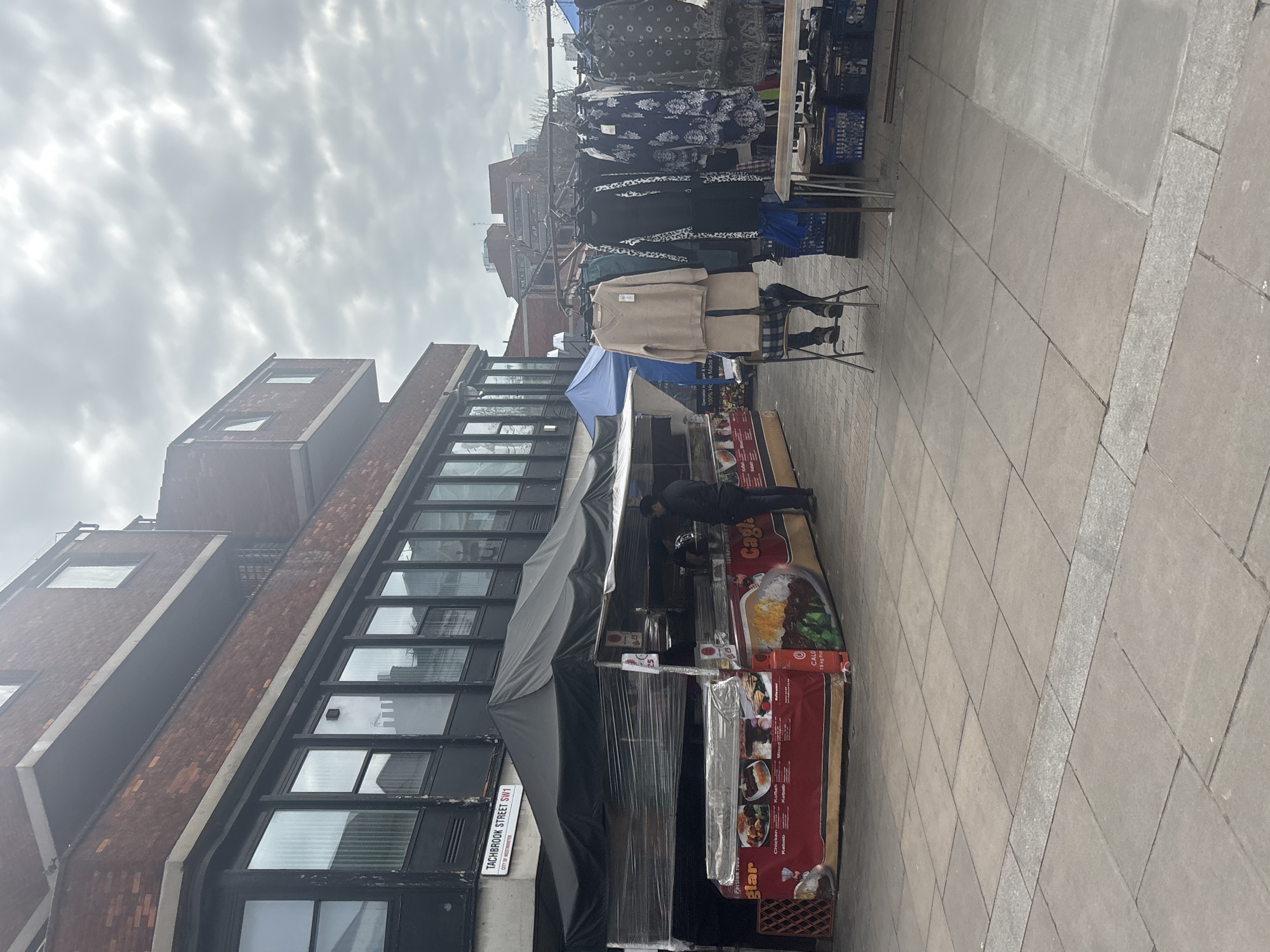 The image shows a street scene in London, United Kingdom, on Tachbrook Street SW1. A food stall is present, advertising “Cagla” with a focus on kebab dishes and "100% Home Made Food”. A person in dark clothing is tending to the stall. To the left is a building with multiple windows and a red-brick façade. Further up the street is a clothing stall with garments displayed on hangers. The sidewalk is paved with rectangular stones. The sky is overcast.