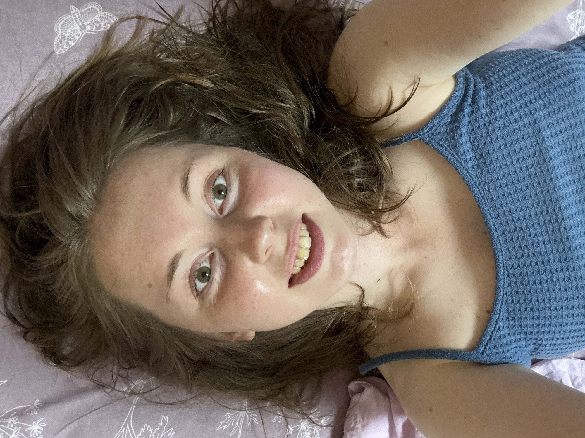 A young woman with brown hair and green eyes is lying on her back, looking up at the camera. She is wearing a blue tank top. Her hair is spread out around her head on a lavender pillow with white butterfly designs. She is smiling, revealing slightly yellowed teeth. The lighting suggests a natural light source, likely from a window. The image appears to be a selfie taken indoors.