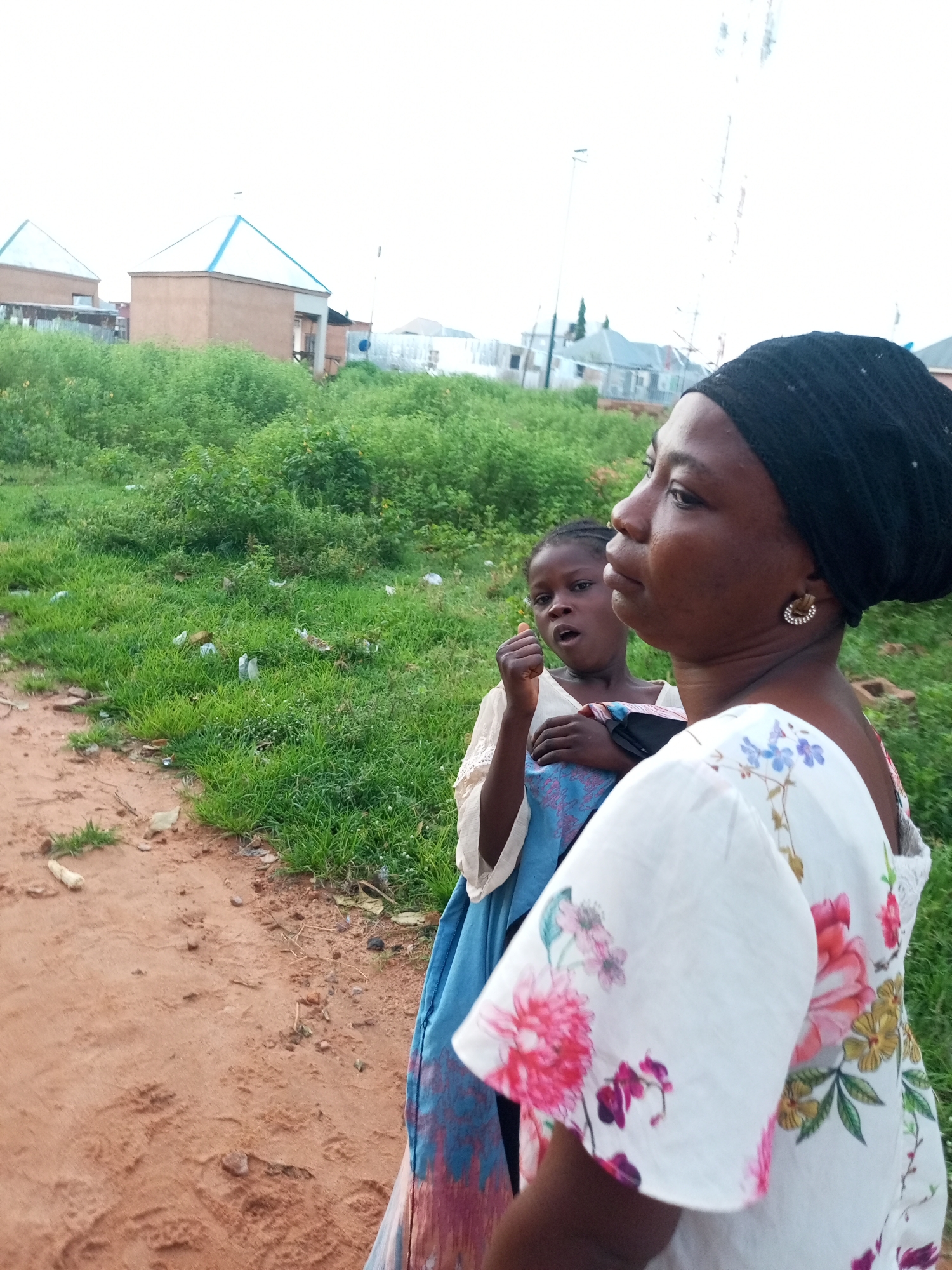 In Tempure, Nigeria, a woman wearing a black headscarf and a white dress with floral print is pictured. She has a small earring in her right ear. A young girl, who appears to be wearing a blue and pink patterned garment, is standing behind her. The two subjects are outdoors, with a dirt path in the foreground and a grassy area with scattered debris in the background. Buildings with peaked roofs and some fencing are visible in the distance. The sky is overcast.