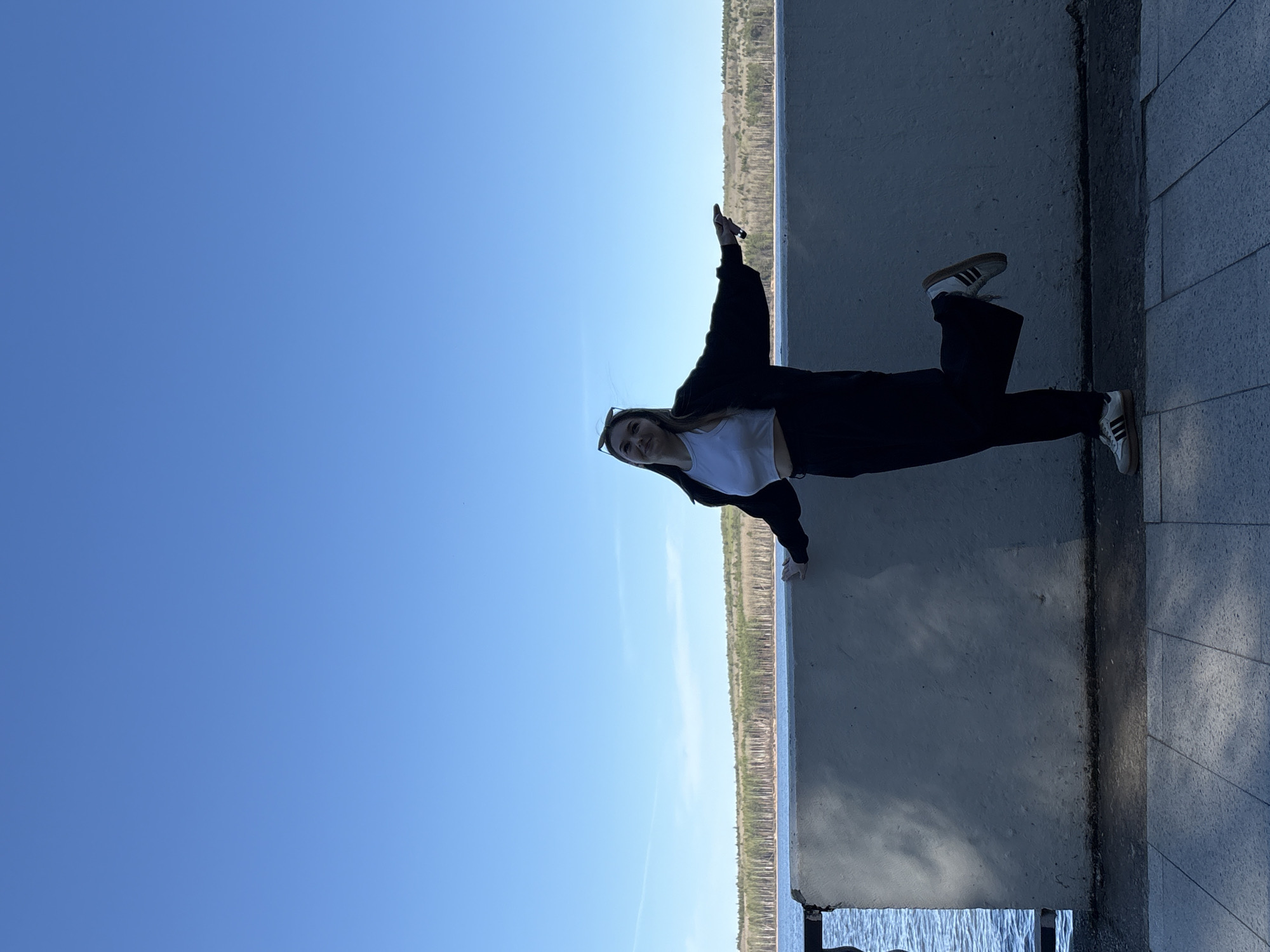 A woman stands on a concrete structure in what appears to be Chaikovsky, Russia, evidenced by the presence of the Kama River and surrounding terrain, in a photograph that is oriented with the subject tilted 90 degrees counterclockwise. She is positioned against a concrete barrier, with her feet planted on a walkway constructed of rectangular stone pavers. The woman is wearing white sneakers with brown stripes, dark trousers, a white top, and a dark jacket. She has one arm raised toward the sky and is smiling. Behind the concrete barrier, a body of water is visible with tree-lined shores in the background. Above this scene, the sky is a clear, pale blue.