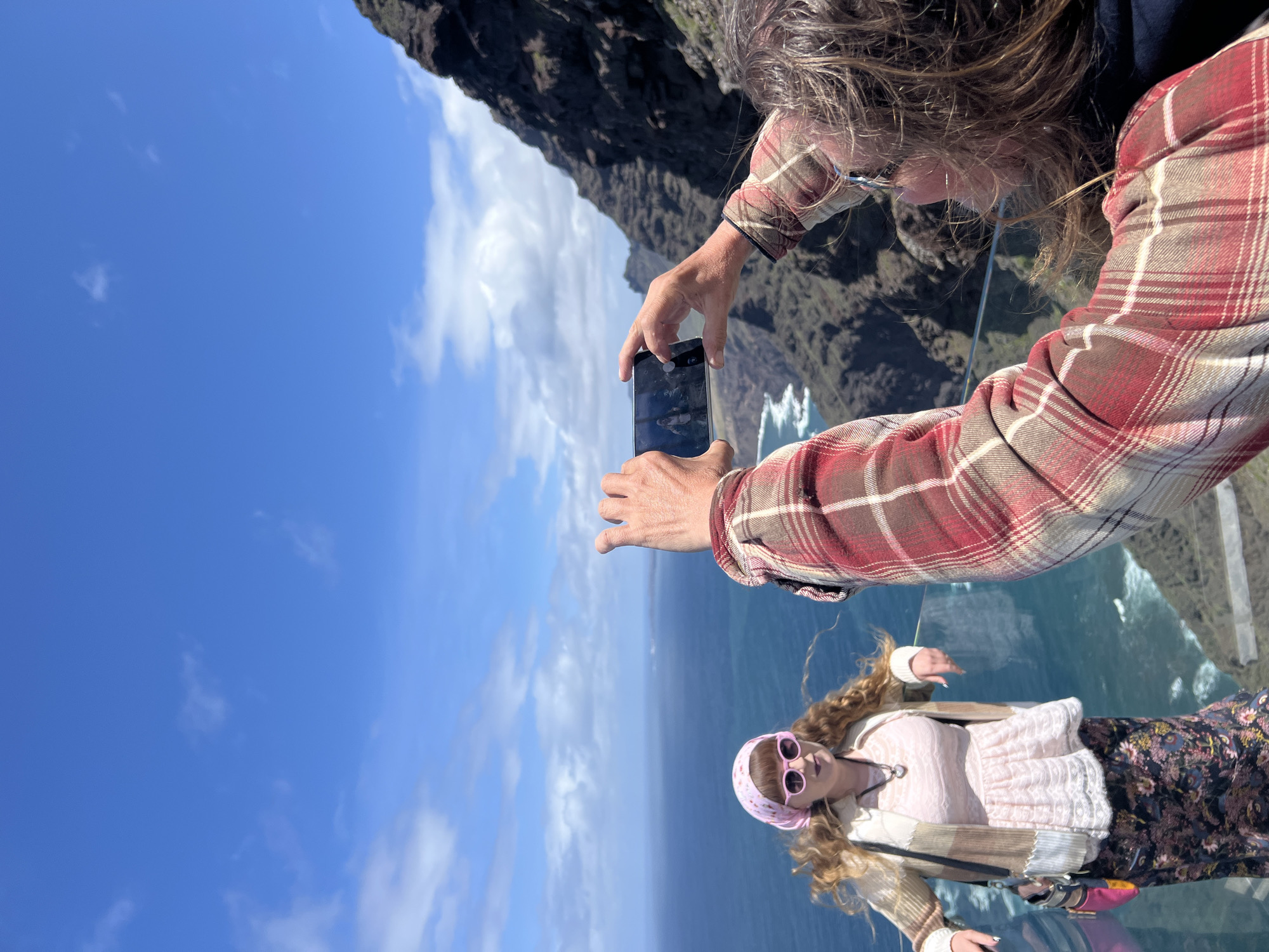 A medium shot captures two individuals: a person photographing a woman on a glass platform. The person taking the photo is positioned higher in the frame and wears a red and beige plaid shirt. They are holding a smartphone to capture the image. The woman being photographed is dressed in a pink top, patterned floral skirt, and a pink patterned headband. She is wearing sunglasses. The photograph is taken at the Mirador del Balcón, a glass platform extending from a cliff in Tirma, Spain. The ocean and blue sky with scattered clouds are visible in the background. A mountainous landscape is also visible behind the photographer.