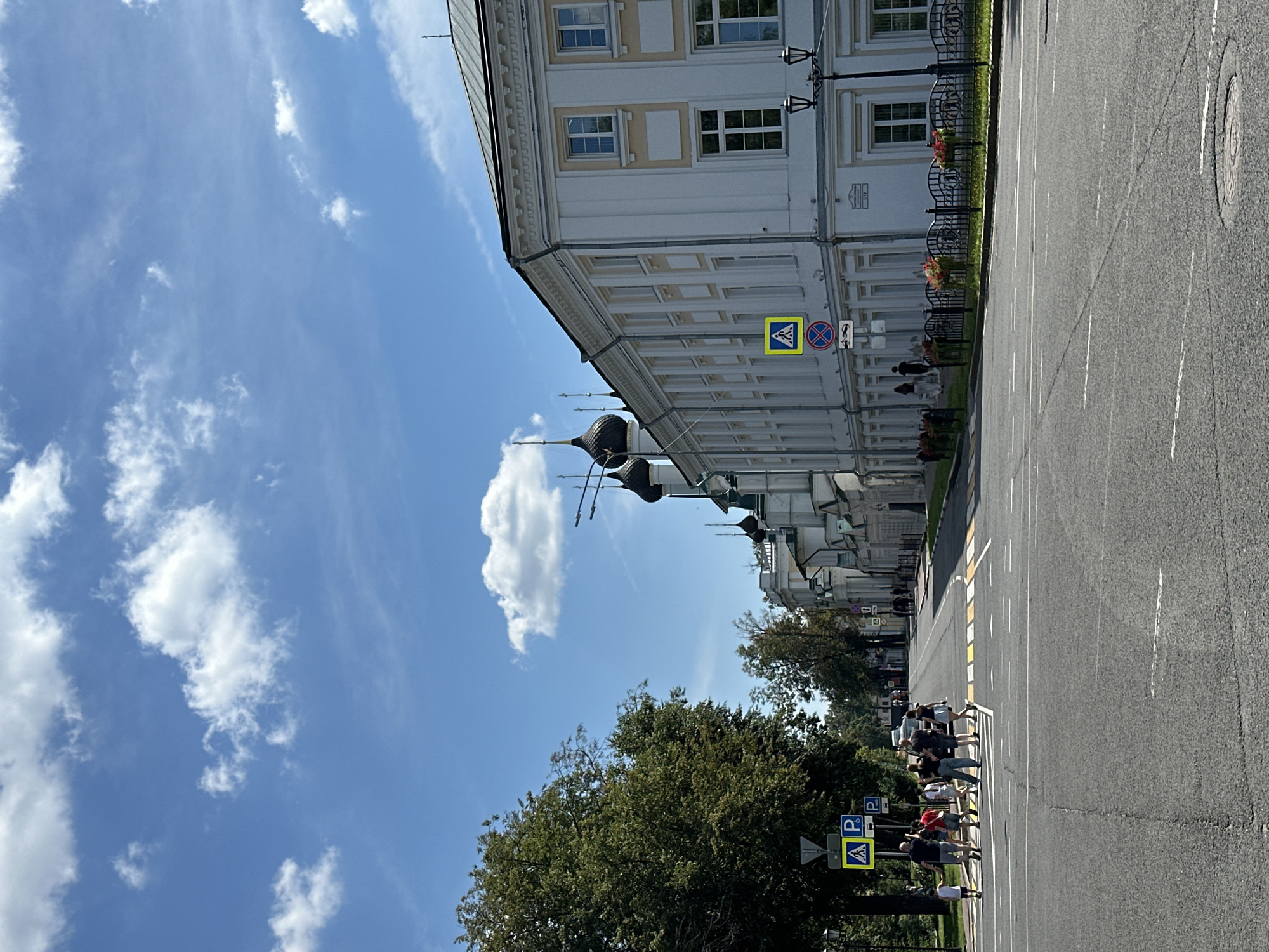A group of seven pedestrians, including children, are crossing a paved street at a marked crosswalk under a clear blue sky with scattered clouds. The street is lined with trees on the left and a long, white building with classical architectural details on the right. The building has multiple windows and a dark, sloped roof. Several traffic signs are visible near the crosswalk, including a pedestrian crossing sign and parking signs. In the distance, more buildings and trees are visible, along with the dark domes of what appear to be churches. The scene is brightly lit by sunlight.