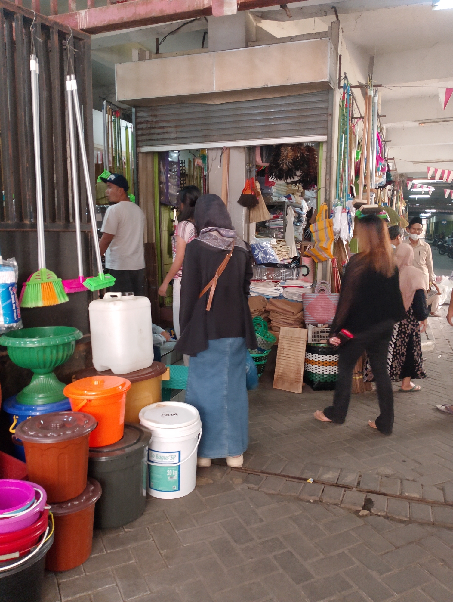 The image depicts a street-level view of a market stall in Magelang, Indonesia. In the foreground, several colorful plastic buckets and containers are stacked, including orange, brown, and green ones. A large white plastic drum is also present. To the left, brooms with green and orange bristles hang from a metal structure. Several people are visible in the scene. A man wearing a white t-shirt and a dark baseball cap stands near the brooms. Further back, a woman in a black hijab and a long blue skirt walks away from the camera. Another woman in a pink and white striped top is partially visible. On the right side of the image, a woman with long brown hair in a black top and pants walks towards the right. A man in a light-colored shirt and a cap, and a woman in a pink hijab and patterned skirt are also present. The background shows the interior of a shop with shelves stocked with various items, including textiles and bags. The ground is paved with brick-like material. The overall scene suggests a busy market environment.