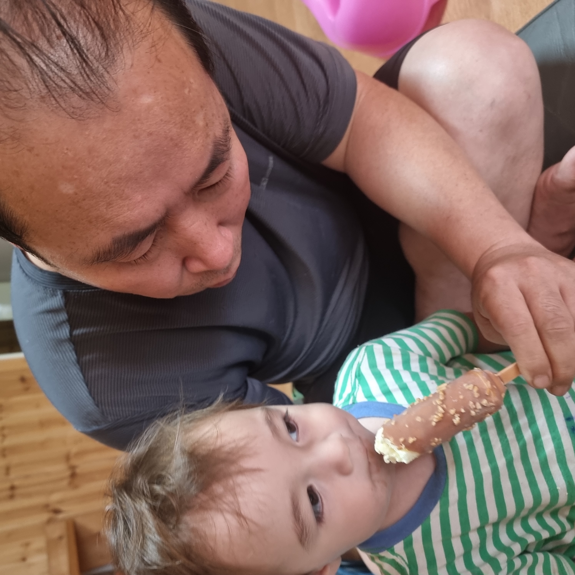 In Dunnae-myeon, South Korea, a child wearing a green and white striped shirt is eating an ice cream bar covered in nuts and chocolate. An adult male, wearing a dark grey t-shirt, is seated and looking down at the child. The background shows a wooden wall.