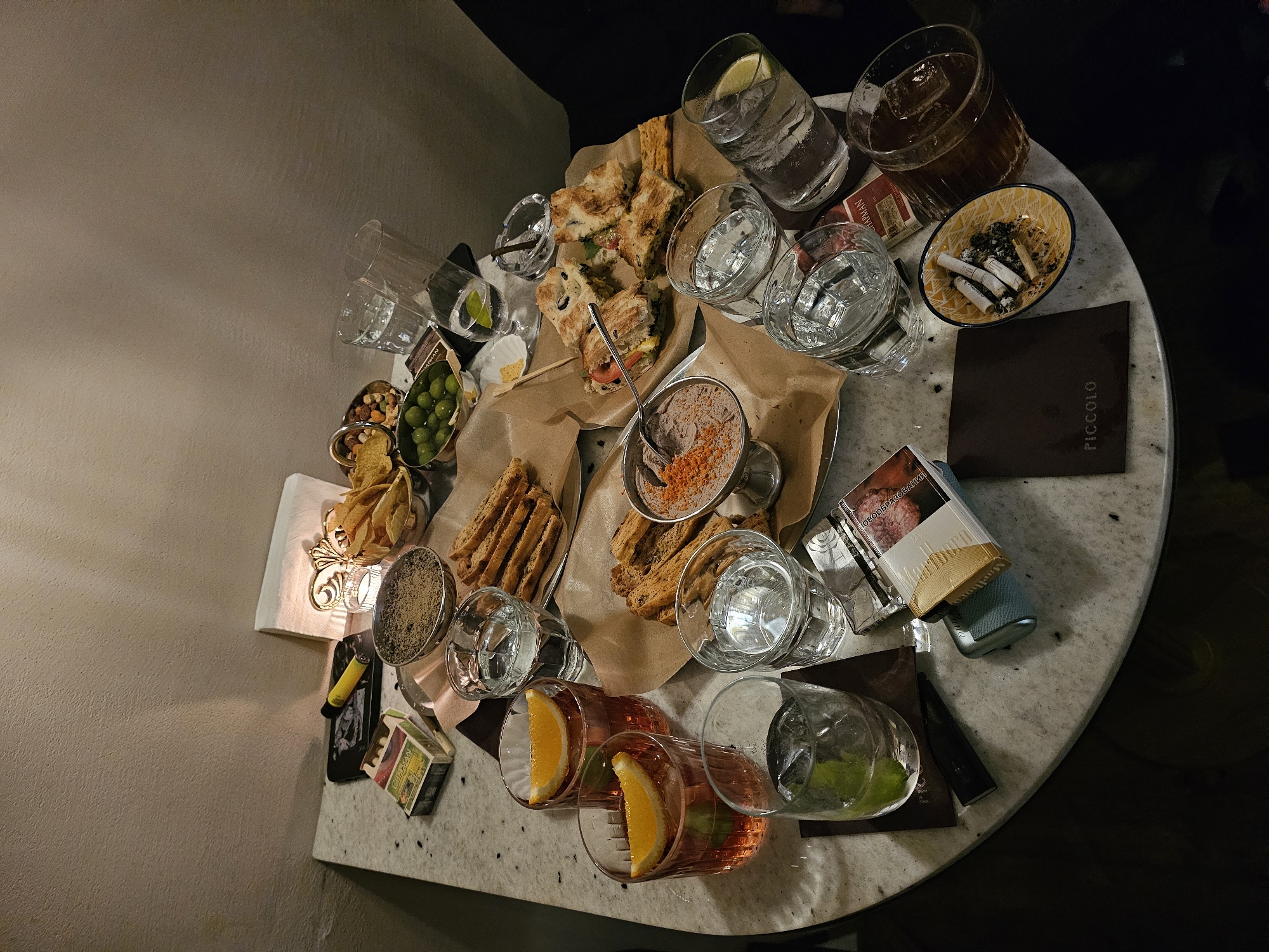A high-angle shot shows a round marble-topped table laden with various food and drink items. The scene suggests a social gathering.
Several drinks are present: glasses filled with clear liquid, some with lemon or lime wedges; two glasses with orange slices, likely spritzes; and a tumbler with a dark liquid and ice, possibly whiskey or a similar spirit. There are several other clear glasses that appear to be empty.
Food items include sandwiches cut into squares, served on brown paper, a small bowl of pate with reddish spices, breadsticks, and snacks like olives, nuts, and chips served in silver bowls.
Various items are scattered on the table, including cigarette packs, a lighter, a mobile phone, and an ashtray containing cigarette butts. A dark brown menu card labeled "PICCOLO" is visible.
The lighting is soft and warm, emanating from a small lamp on the left of the table, suggesting a relaxed and intimate setting. The scene is located in the city of Unknown, Unknown. The environment is dimly lit.