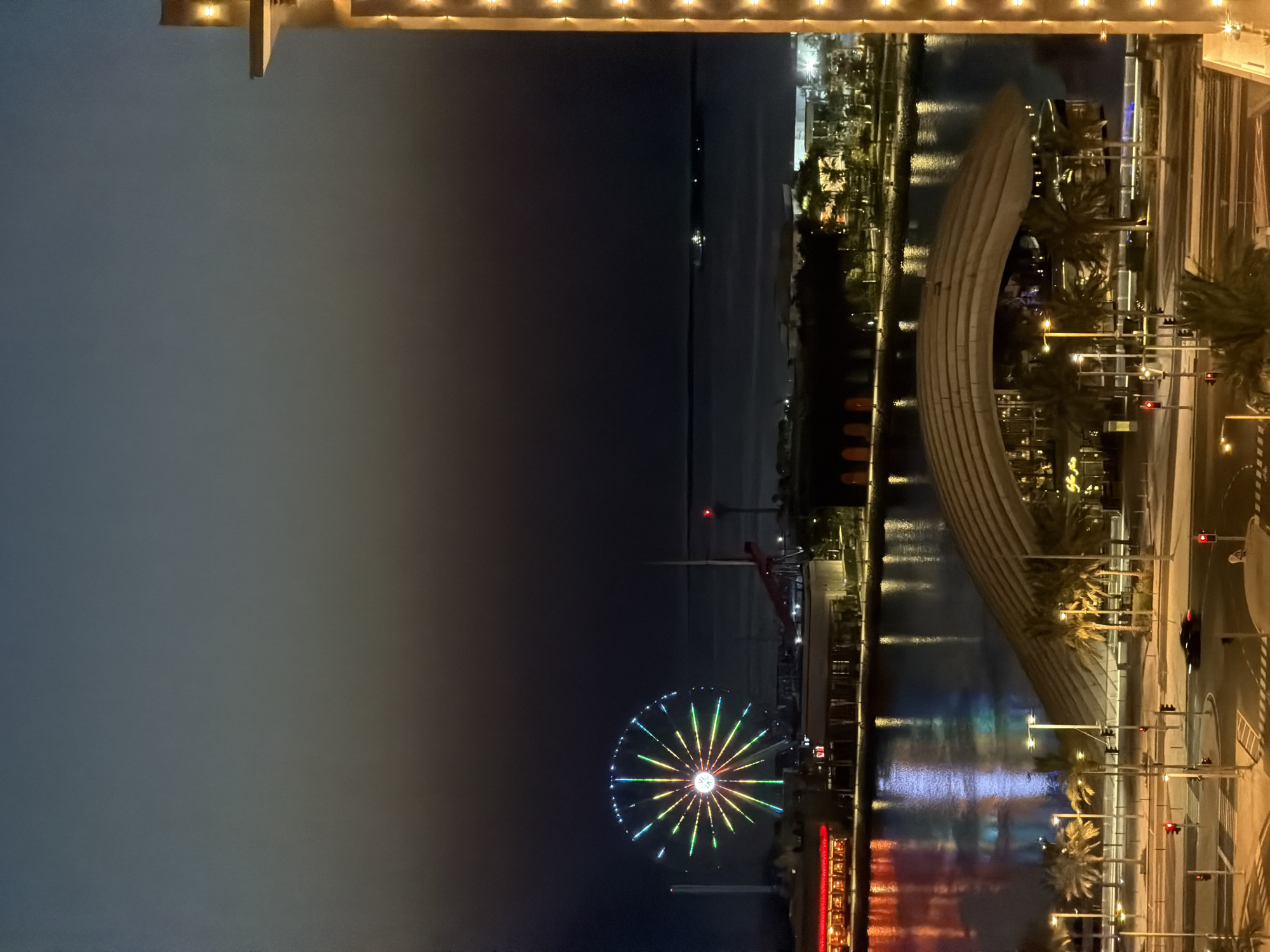The image depicts a nighttime scene of a waterfront area in an unknown city. Dominating the left side of the frame is a brightly lit Ferris wheel, displaying a vibrant array of colorful lights. Adjacent to the Ferris wheel are buildings with illuminated windows, reflecting in the dark water of a canal or bay. In the foreground, a modern architectural structure with a curved roof is visible, surrounded by palm trees and streetlights. A street with traffic lights and a moving vehicle is also present. The sky is a gradient of dark blue and gray, suggesting dusk or early night. On the right side of the image, a portion of a building or structure with decorative lights is in view. The overall atmosphere is one of urban entertainment and nighttime activity.