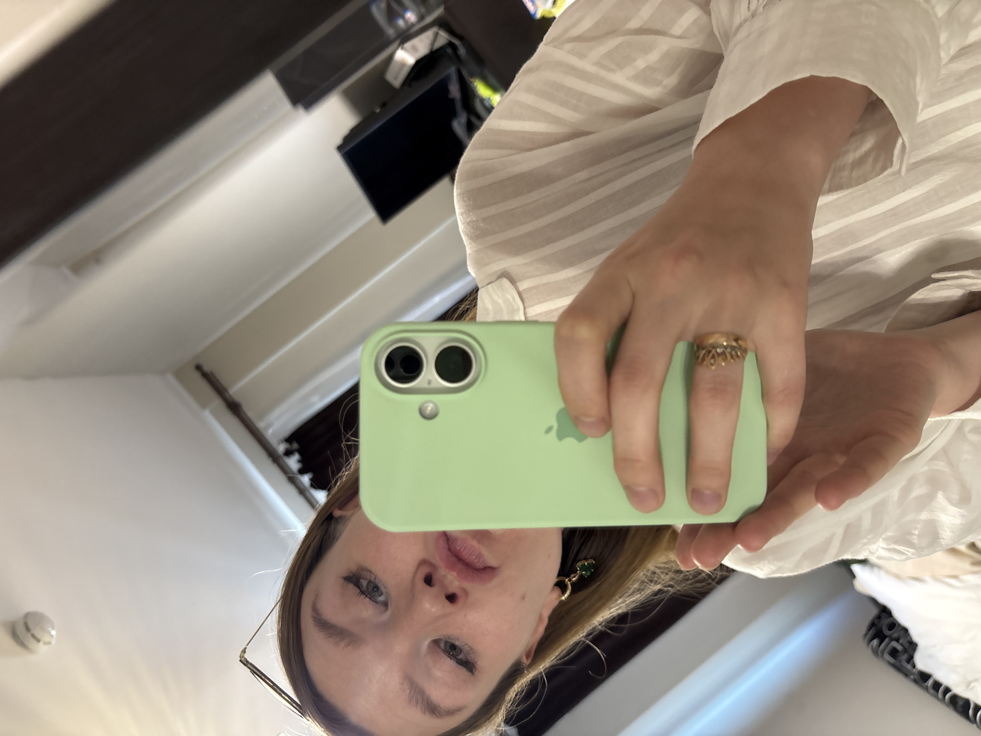 The image shows a young woman taking a selfie in what appears to be a hotel room. She is holding up a light green iPhone in her right hand, angled towards the mirror. She has light brown hair, is wearing a white striped shirt, and has subtle makeup. She is also wearing earrings and a ring on her left hand. The background shows a mirror reflecting the room, with hints of furniture, a dark computer monitor, and a white ceiling with a smoke detector. The context of Izhevsk, Russia, cannot be determined from the visual information in the image.