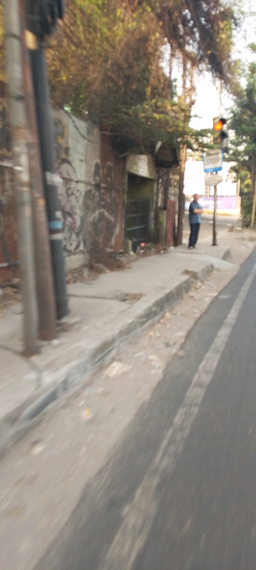A person stands at a bus stop on a sidewalk next to a street in Bekasi, Indonesia. The person is wearing a dark blue shirt and dark pants. They are standing with their arms crossed and looking away from the camera. Behind the person, a bus stop sign reads "STOP". A traffic light is visible to the right of the bus stop sign, with the red light illuminated. The street appears to be paved and there is a sidewalk bordering it. A wall with graffiti can be seen to the left of the sidewalk. Trees and foliage are visible in the background, suggesting an outdoor urban environment. The overall scene is somewhat blurry, indicating motion during the capture of the image.