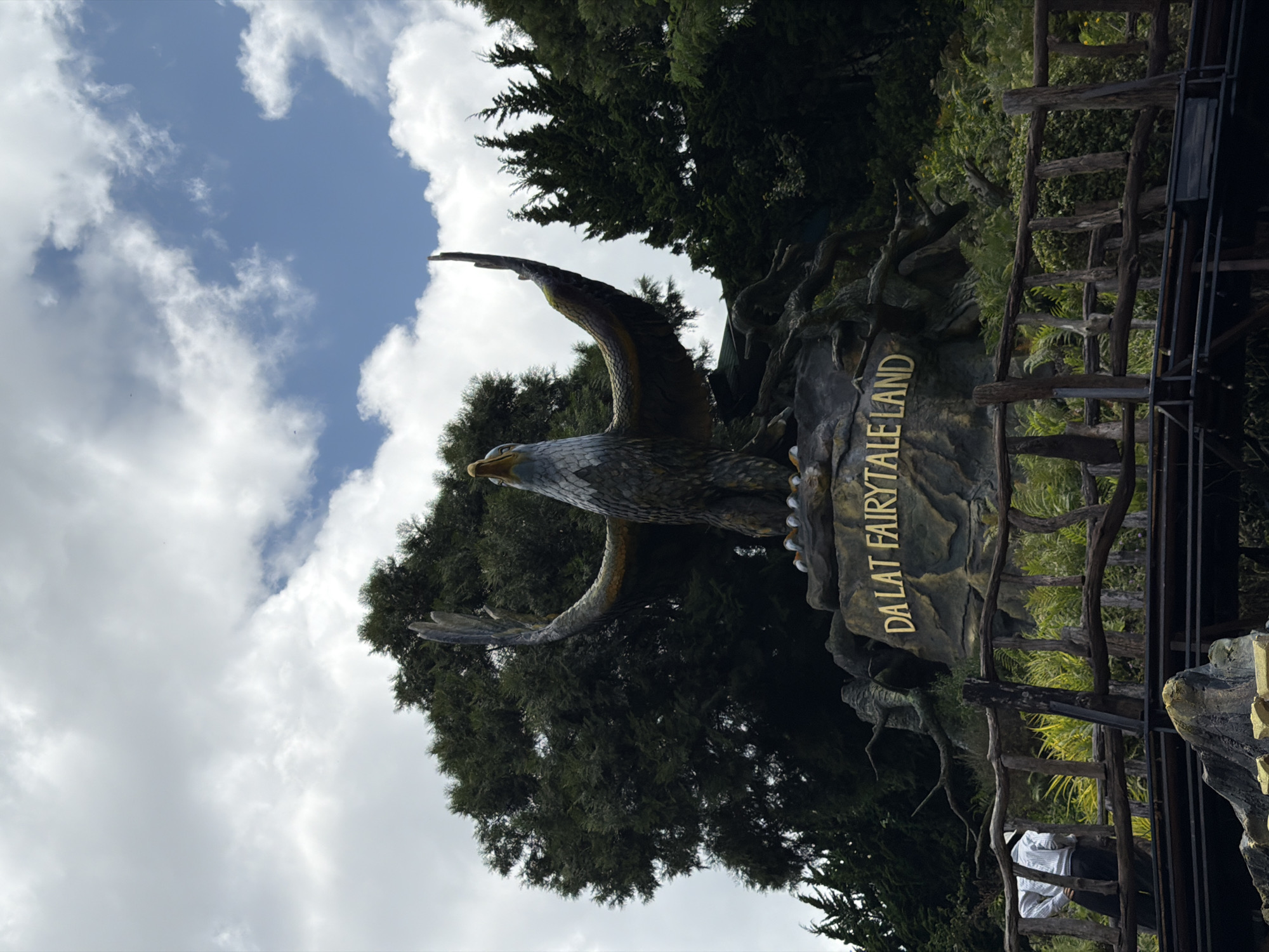 The image shows a large eagle statue with wings spread wide, positioned on a rocky outcrop. The statue is located at "Dalat Fairytale Land" in Da Lat, Vietnam, as indicated by golden lettering on the rock. Behind the statue, a dense forest of dark green trees forms the backdrop, with a cloudy sky overhead. In the foreground, a wooden fence and railing structure with a bridge or walkway can be seen. One person, wearing a white shirt and dark pants, is partially visible on the left side of the fence, looking away from the camera. The overall scene suggests an outdoor attraction or park.
