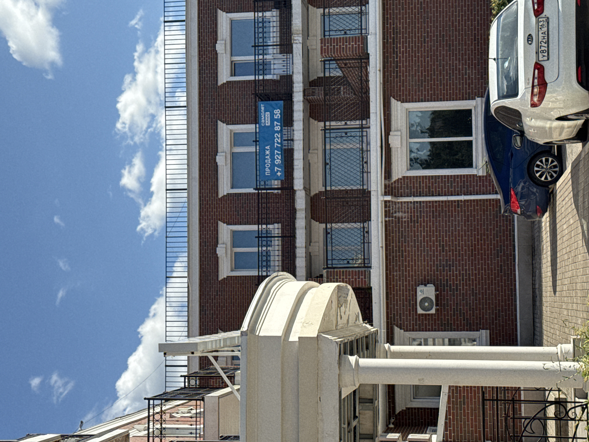 The image depicts a modern brick building with multiple floors and balconies. The sky is blue with scattered white clouds. In the foreground, there is a paved area with two cars parked, one dark blue and one white. The white car has a license plate that reads "Y872HA 163". On the second floor of the building, a blue banner with white text is displayed on a balcony, which appears to be an advertisement for a sale. The building has several windows and air conditioning units mounted on the exterior walls. There is also a decorative stone structure with pillars in the left foreground, suggesting an architectural element of the building or its surroundings.