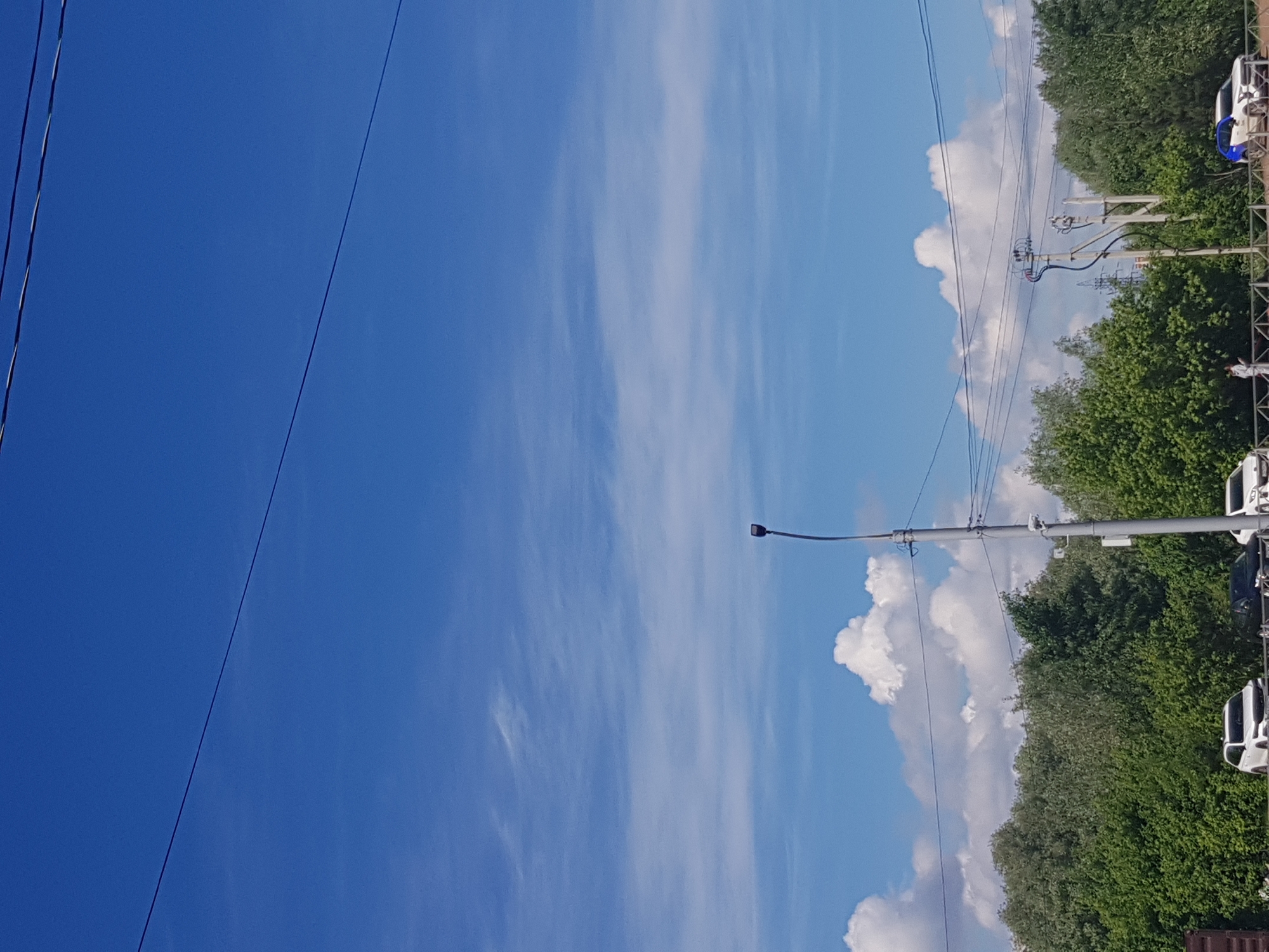 The image shows a street scene under a bright blue sky with streaky cirrus clouds. Power lines run diagonally across the frame from the top left to the center right. A street lamp is visible on the right side of the image. Green trees line the background on the right, behind which there are power lines and cars are parked. The location is Unknown, Unknown.