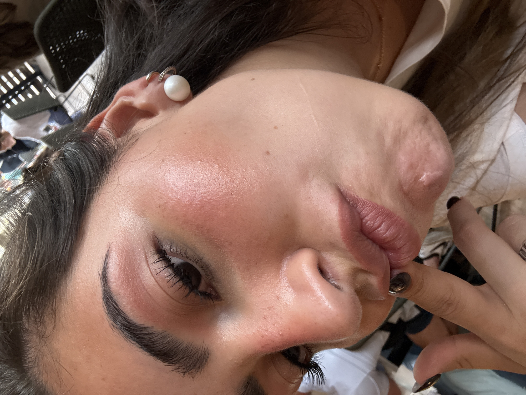 A close-up, low-angle shot depicts a woman with dark hair, fair skin, and prominent facial features, likely in an outdoor setting. She has a pearl earring in her left ear, defined eyebrows, and visible eyelashes. Her lips are slightly parted. A finger with dark-polished nails is visible near her mouth. The background is somewhat blurred but appears to include objects like chairs and other people, suggesting a public or semi-public outdoor space. Without additional context, determining the precise location in San Antonio is impossible.