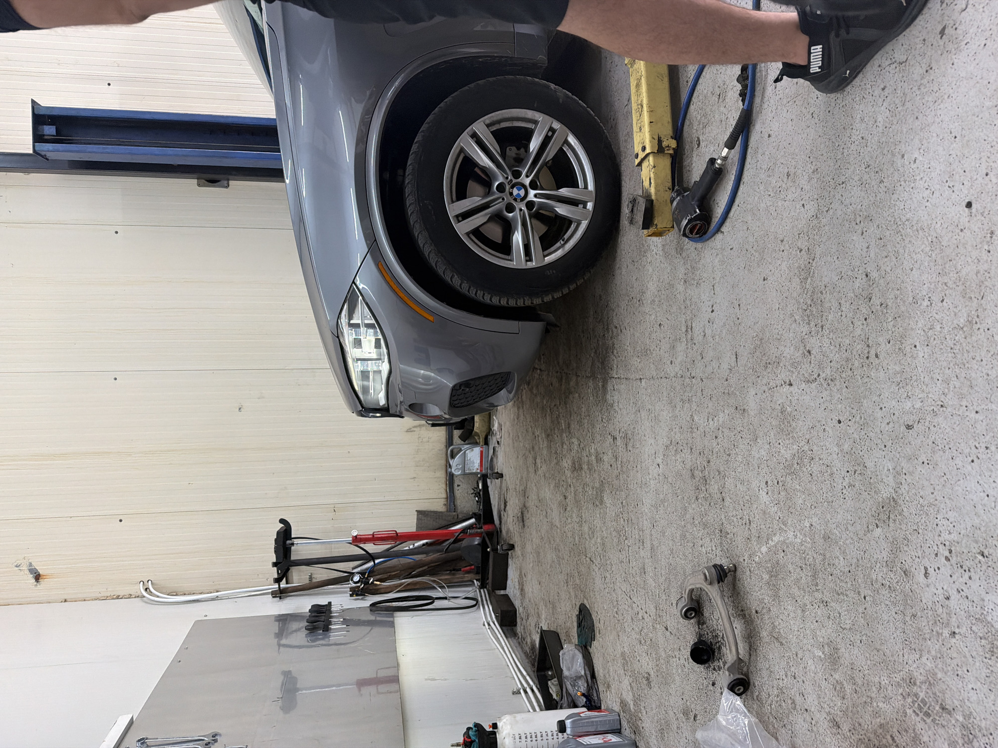 This is a forensic, matter-of-fact description of an image taken in a workshop located in Sofia, Bulgaria. The scene depicts a gray BMW SUV parked on a concrete floor. To the left, a metal cabinet with tools mounted on its surface is visible. Various car maintenance equipment, including a red hydraulic jack and other tools, are scattered around the vehicle. A detached car suspension component lies on the floor in the foreground, near a plastic bag. A person, partially visible from the waist down, is standing to the right of the frame, wearing black Puma sneakers. The workshop has ribbed, light-colored walls and a blue car lift in the background. The overall environment suggests an active automotive repair setting.