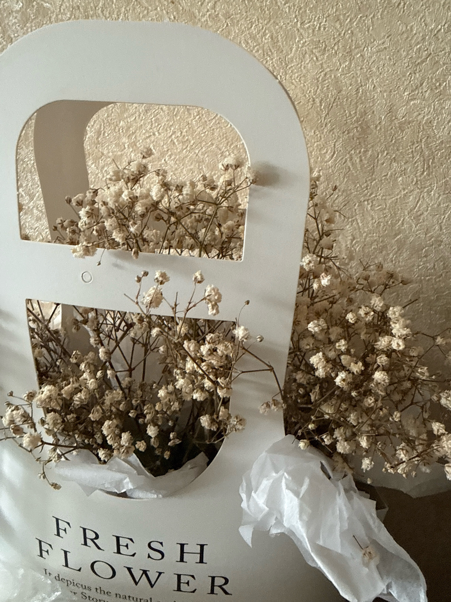The image shows a bouquet of dried baby's breath flowers in a white paper gift box. The box has a cut-out handle and the words "FRESH FLOWER" printed on it. The background is a textured beige wall. The lighting is soft and natural, suggesting an indoor setting. The specific location in Zavolzhye, Russia, is not discernible from the image.