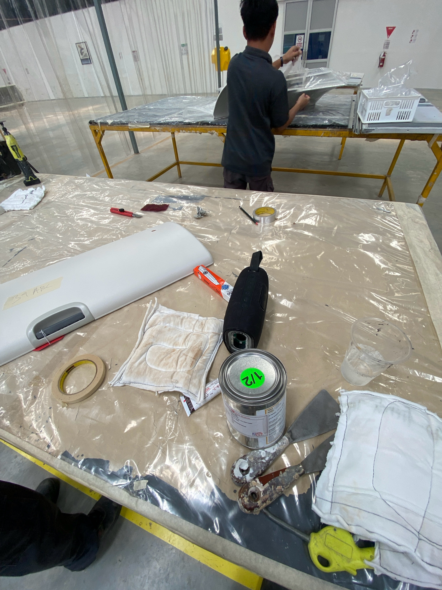 A man is working on a large white object on a table covered in clear plastic sheeting, in what appears to be a workshop or industrial space in Batam, Indonesia. The man is wearing a dark grey t-shirt and dark shorts and is seen from behind as he handles a large piece of grey material, possibly part of the white object, which is resting on another table covered in plastic.
In the foreground, the white object, which resembles a section of a fuselage or a large curved component, has a label with handwritten text "34 ABC". Several tools and materials are laid out on the plastic sheeting around the white object. These include a roll of masking tape, a red utility knife with a maroon scrubbing pad nearby, a black portable speaker, a can of what appears to be sealant or resin with a green sticker labeled "1/2", a clear plastic cup containing a clear liquid, two metal spatulas with worn handles, and several white cloths, one of which is stained. A yellow industrial tool, possibly a router or grinder, is also visible in the lower right corner, partially covered by a white cloth.
The environment seems to be an indoor facility with high ceilings and exposed structural elements. The background shows industrial shelving and what appears to be a partition made of vertical plastic sheeting. Natural light is entering the space, creating reflections on the plastic surfaces. The floor is concrete.