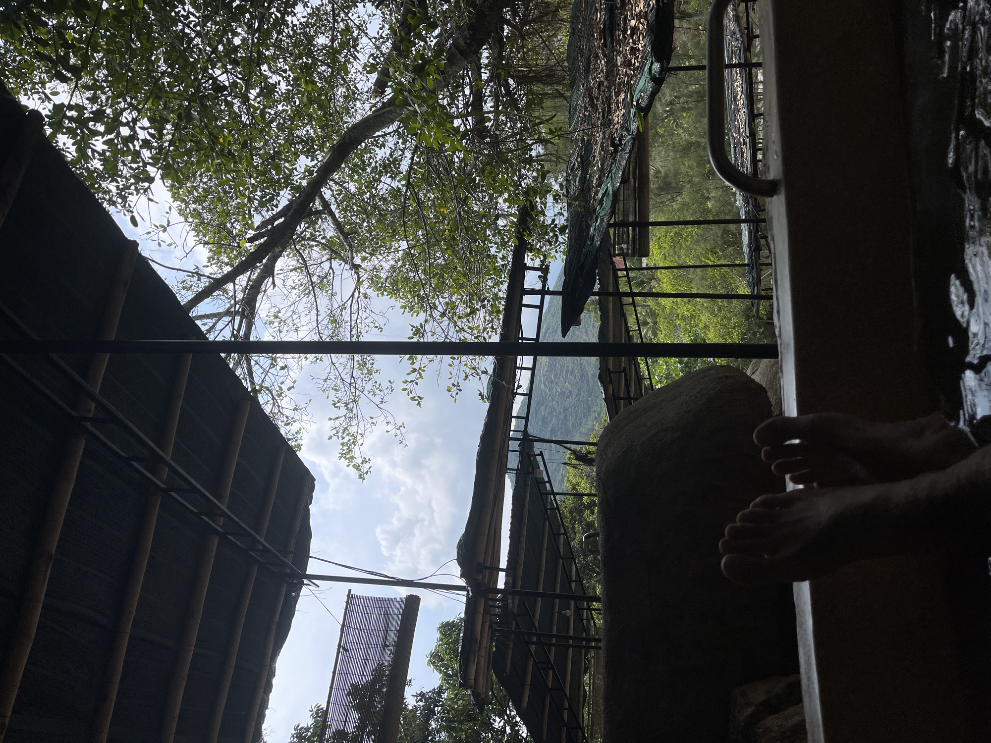 The image shows a partial view of a hot spring or bath area. From the perspective of someone inside the bath, the view includes two feet immersed in the water, and a stone border to the left. Beyond the border, a landscape includes trees and vegetation. Above, a structure of bamboo and metal scaffolding is visible, suggesting a roof or enclosure, along with the sky with clouds. The foliage of nearby trees is also present. The image is rotated, making the composition seem tilted. Due to the provided information, it's assumed that the location is likely a hot spring area within or near the city of Tây Nha Trang, Vietnam.