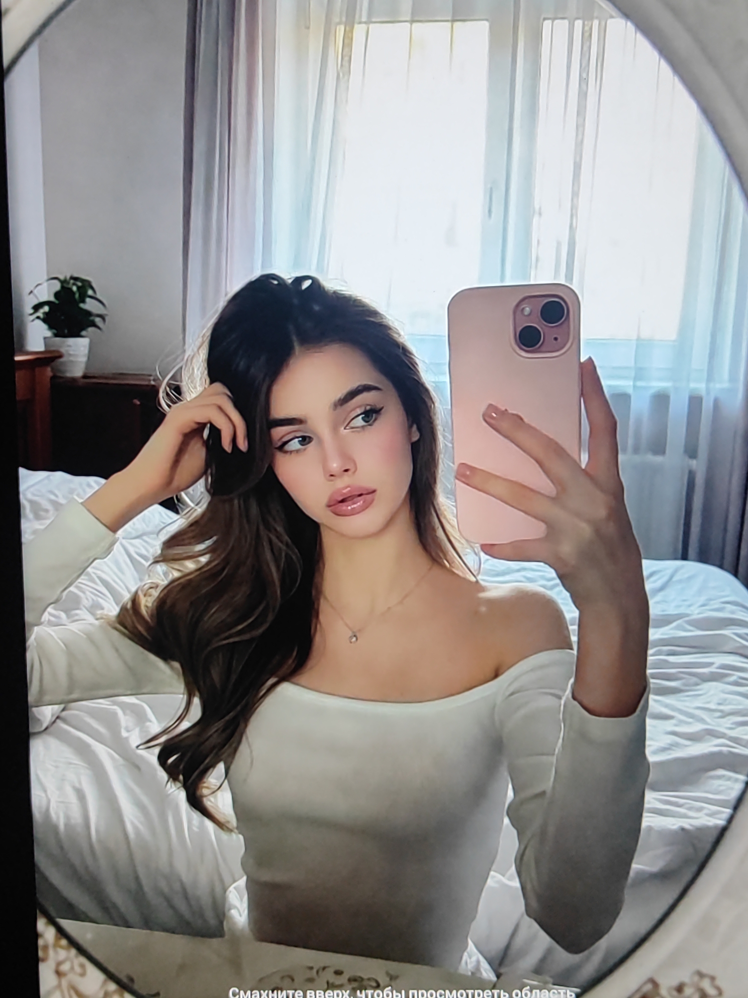 This is a selfie of a young woman taken in a bedroom. She is leaning against a bed covered in white sheets, with her hair falling over her shoulder. She is wearing an off-the-shoulder white top and has a delicate necklace with a small pendant. She is holding a pink smartphone in her right hand, which is capturing the reflection. The woman has long, dark, wavy hair and is looking off to the side with a slight smile. Her makeup is natural, with emphasis on her eyes and lips.
In the background, sheer curtains cover a window, allowing soft natural light to filter into the room, suggesting it is daytime. To the left, a dark wooden dresser with a potted plant on top is visible. The overall atmosphere is relaxed and intimate, likely a personal moment captured at home.
The image contains visible text at the bottom which reads "Смахните вверх, чтобы просмотреть область". This text is in Russian and translates to "Swipe up to view the area".