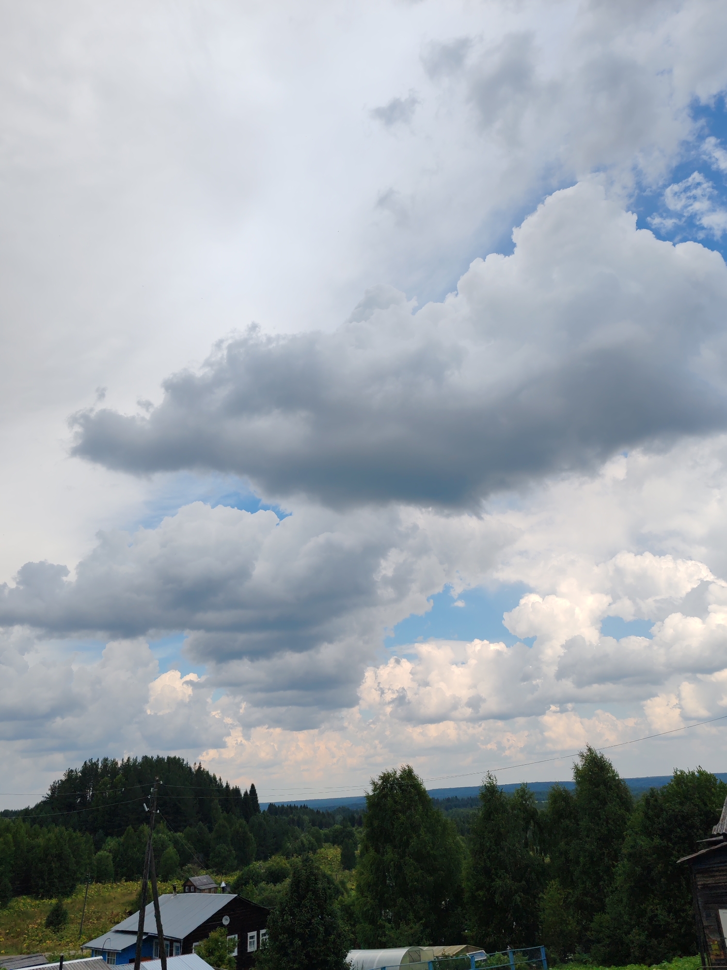 The image captures a rural landscape under a cloudy sky, likely in or around Чухлэм, Russia. The foreground features a dark wooden house with white-trimmed windows and a dark, gabled roof. To the left of this house, a smaller blue and white building is partially visible. A power pole with wires stands prominently in the middle ground, to the left of the houses.
Beyond the structures, a dense forest covers a gently sloping hill. Further in the distance, a rolling landscape of trees stretches towards the horizon, under a dramatic sky filled with large, volumetric white and gray clouds interspersed with patches of blue sky. The overall impression is one of a serene, rural setting with signs of habitation within a natural environment.