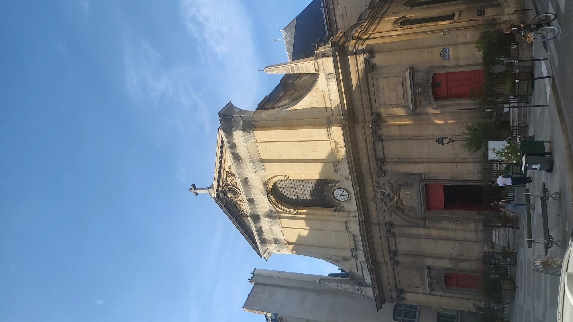 A wide-angle, eye-level shot captures the facade of the Saint-Roch Church in Paris, France, under a bright blue sky with scattered wispy clouds. The church, constructed of light-colored stone, features classical architectural elements. A prominent cross tops the pediment, which is adorned with intricate carvings. Below the pediment, a large arched window with stained glass is visible, and beneath that, a clock face shows the time as approximately 10:10. The church's entrance is flanked by two red doors, one on each side, and a larger set of red double doors in the center. A street lamp stands to the right of the central entrance. In the foreground, several people are present on a paved square in front of the church. Two individuals, one in a light-colored shirt and the other in a denim jumpsuit, walk towards the church. A woman is riding a bicycle on the right side of the frame, while a bench sits in the middle ground. Planters with greenery are placed around the square. A trash receptacle is visible near the bench. A street sign that reads "PLACE SAINT-ROCH" is affixed to the wall of the church on the right side. The building to the left of the church is a tall, modern structure with windows.