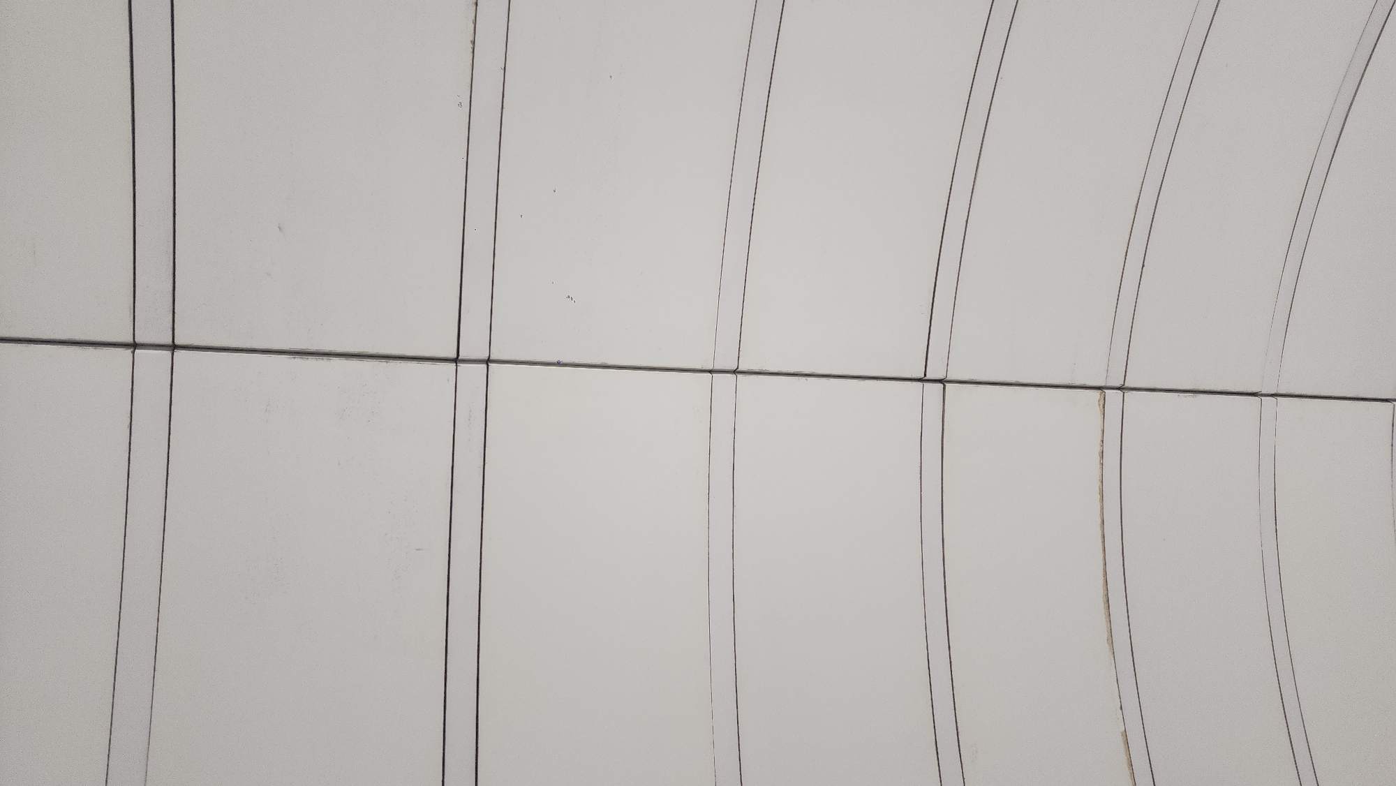 The image shows a section of a curved, white panelled ceiling. The panels are rectangular and arranged in rows. Dark lines delineate the edges of the panels, both horizontally and vertically. The horizontal lines run straight across the image. The vertical lines curve in an arch. There are some minor discolorations and marks on some of the white panels. The location and city are unknown.