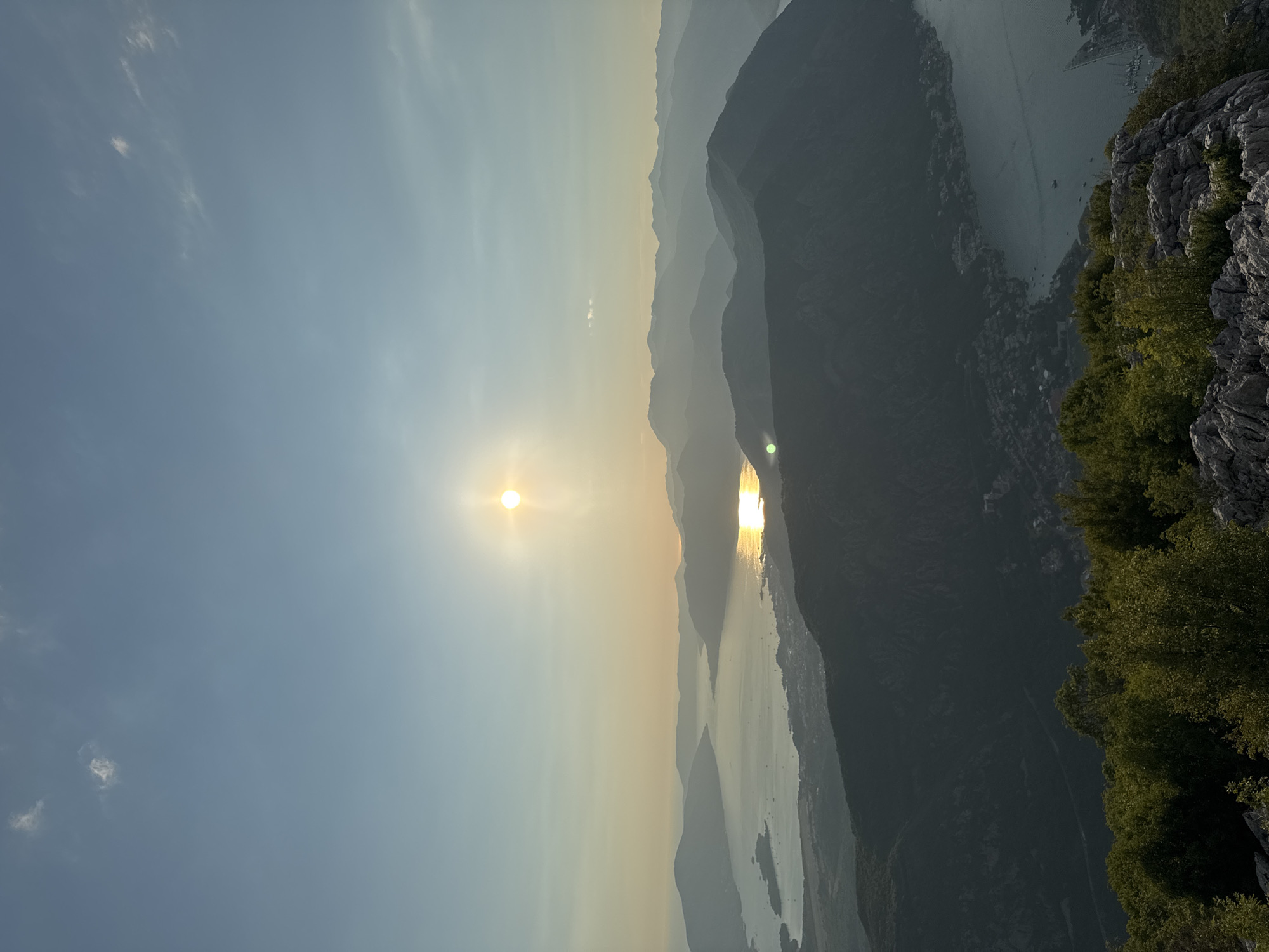 This is a high-angle, wide shot taken from a mountain overlooking a bay. The sun is setting, casting a golden glow on the water and creating a hazy effect over the distant mountains. The foreground is dominated by rugged rocks and lush green trees. In the middle ground, the bay is visible, with small islands and a coastline dotted with buildings. The background consists of layers of silhouetted mountains fading into the horizon. The sky is a clear blue with a few wispy clouds. The image was captured in Žanjev Do, Montenegro.
