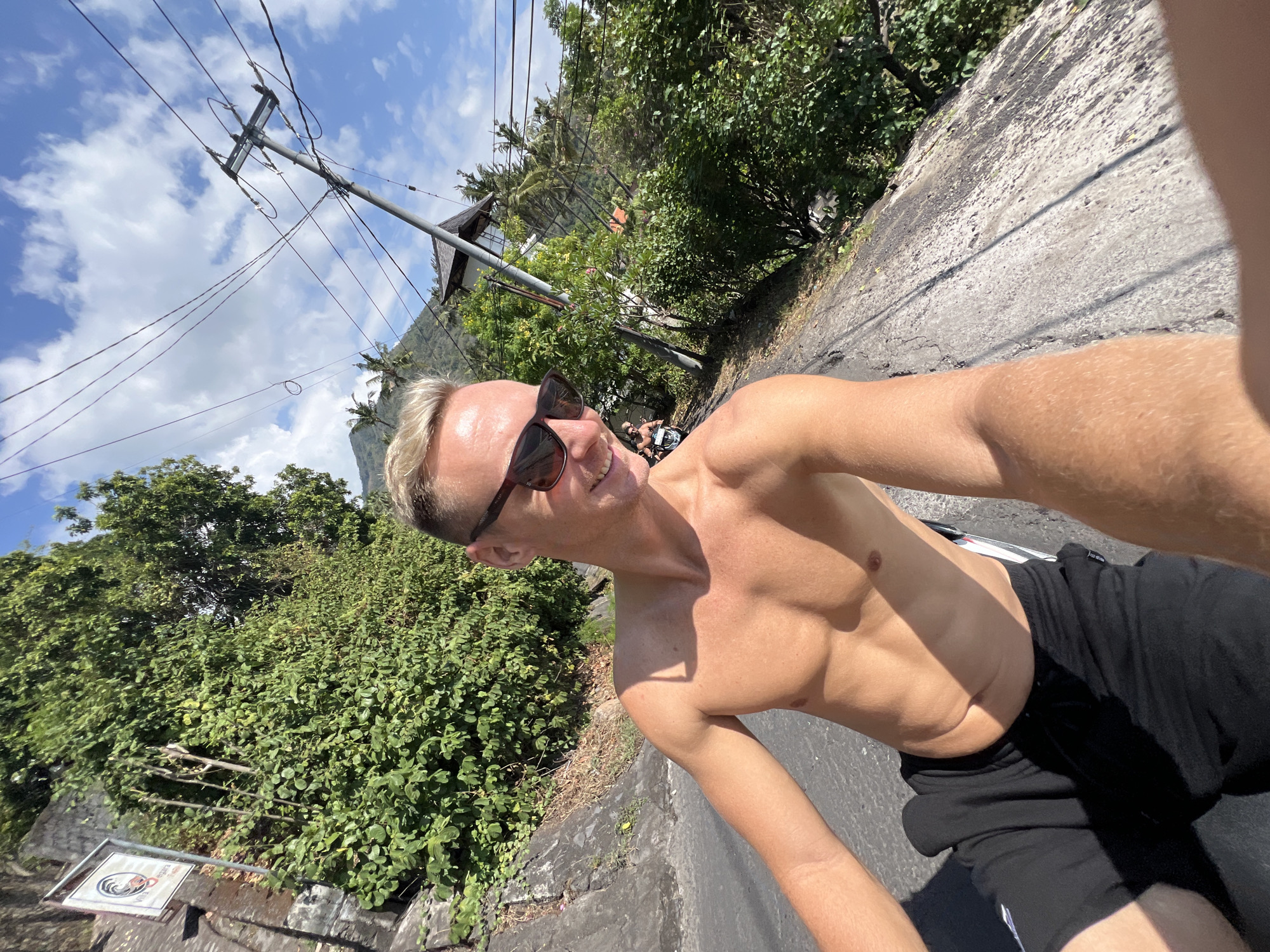 A Caucasian man with blonde hair and sunglasses is riding a motorcycle down a road in Abang, Indonesia. He is shirtless and wearing black shorts. The road is paved and has a steep embankment on the left side covered in lush green foliage and a stone wall. There are power lines overhead and a mountain in the background. Another person is visible riding a motorcycle in front of the subject. The sky is blue with scattered white clouds.