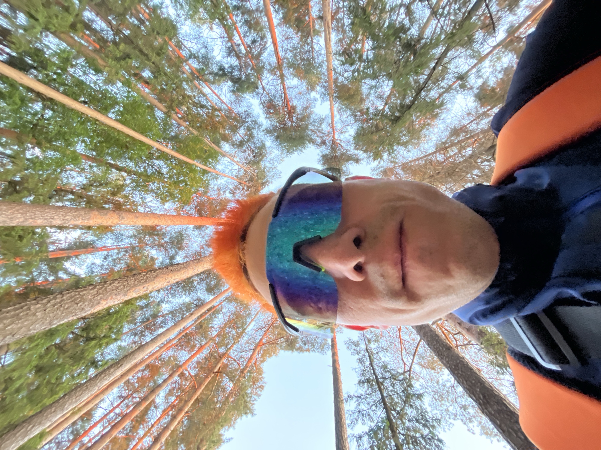 This forensic description details a low-angle self-portrait taken in a conifer forest in Chaikovsky, Russia. The image captures an individual's face, angled sideways from the lower-center to the right, looking upwards towards the sky and tree canopy. The person, an adult with short, reddish-orange hair visible on the left side of the head, wears iridescent, mirrored sports sunglasses that obscure the eyes and upper face. The lower portion of the nose, mouth, and chin are visible, with lips slightly parted. The individual is wearing a dark blue garment, possibly a hoodie or jacket, with a prominent orange strap or band extending horizontally across the chest area from the right.
The background consists of a dense forest of tall, slender conifer trees, likely pines, with reddish-brown trunks and green foliage. The trees extend vertically, appearing to converge towards a central point overhead due to the wide-angle, upward perspective. The sky, visible between the tree crowns, is a clear, light blue. Sunlight appears to illuminate the tree trunks from the side, casting warm tones. The overall scene depicts a person positioned directly beneath a forest canopy, capturing a selfie from a worm's-eye view.