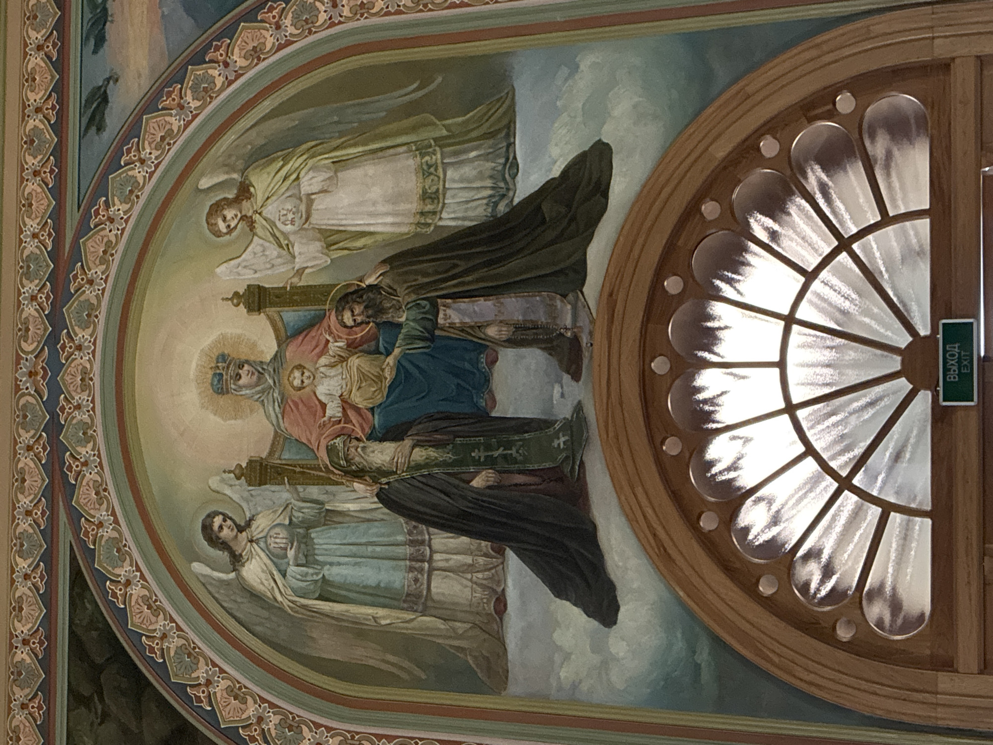 A mural adorns a wall, possibly inside a building in Боголюбово, Russia. The mural is in an arched frame with ornate borders. The scene depicted is a religious one, featuring the Virgin Mary and a young Jesus, attended by angels. Several figures surround them, possibly saints or other religious figures, all within a cloudy, heavenly environment. To the right of the mural is a window with a stylized floral design. An exit sign is present on the window frame, with text in Cyrillic and English.