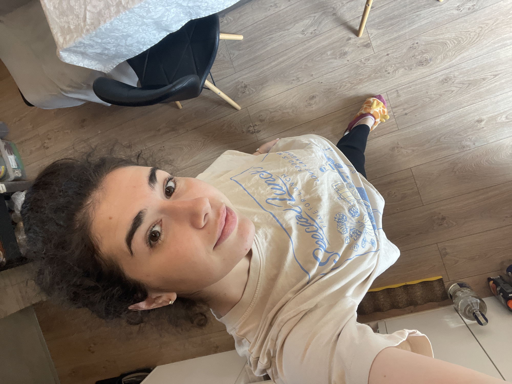A young woman with curly dark hair is positioned in the lower left of the frame, looking up and towards the camera. She is wearing a beige t-shirt with blue graphic text. Her right ear is visible, adorned with a small earring.
The scene includes elements suggesting an interior setting: part of a white tablecloth covering a table, a black chair, and wood-look flooring. Further to the right are objects including a cat scratching post, glass water bottle, and a can of compressed air. At the top-right a leg clad in black pants and wearing a yellow and pink slipper is visible.
There is insufficient information to place the scene in Sofia, Bulgaria.