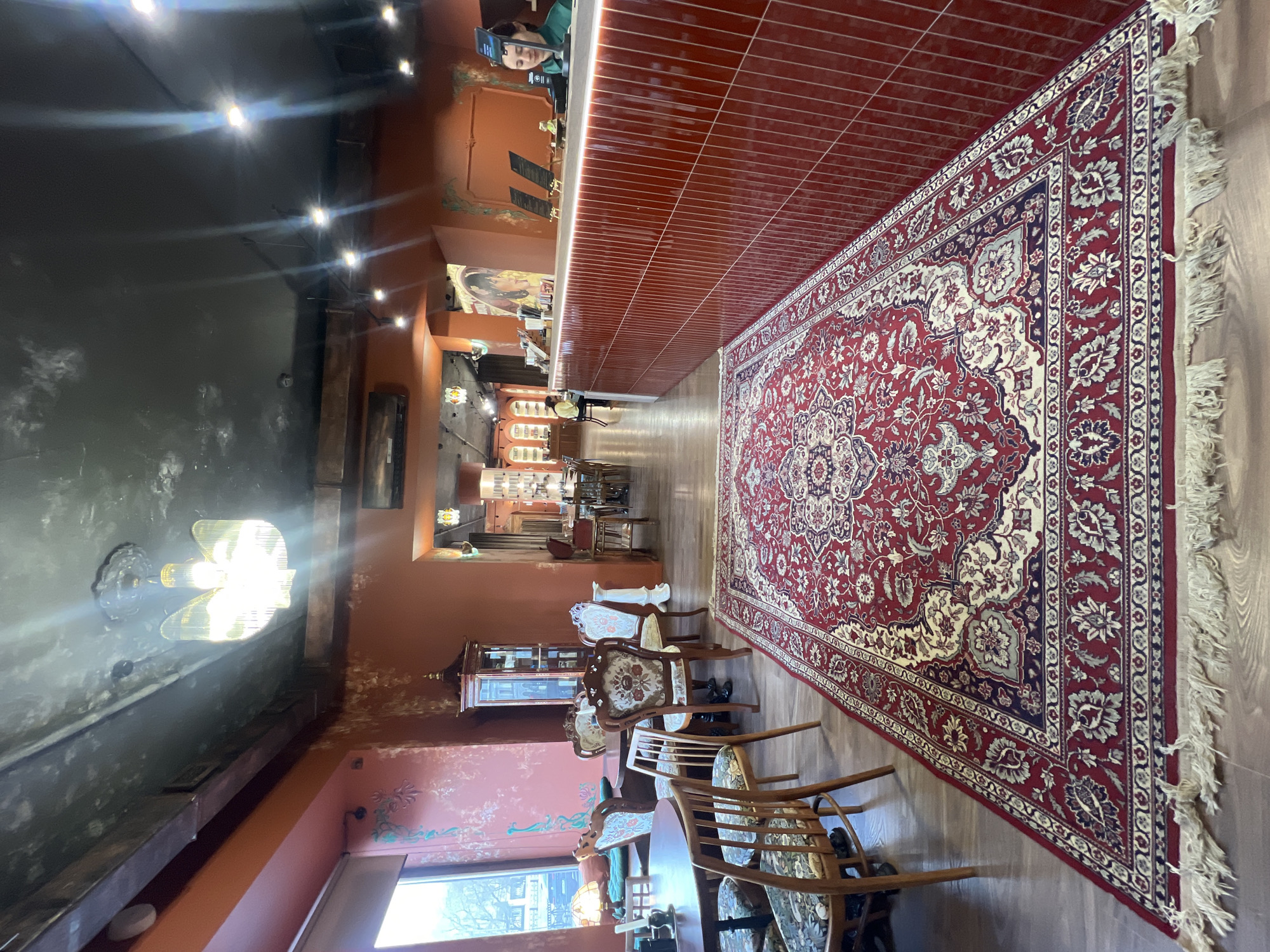 The image displays the interior of a cafe, restaurant, or lounge, characterized by a warm and somewhat dim atmosphere. The perspective looks down a long, central aisle of the establishment.
The floor is light-colored wood, mostly covered by a large, ornate Persian-style rug in shades of red, cream, and dark blue/gray, featuring intricate floral and medallion patterns. The walls are primarily painted in a terracotta or burnt orange color, with some sections on the left exhibiting subtle, decorative patterns. A darker, possibly black or dark gray, wall section with illuminated wall sconces is also visible on the left. The ceiling is dark, equipped with multiple recessed spotlights and a prominent chandelier on the left.
On the right side of the image, a long counter with a light-colored top and a front covered in dark red tiles extends into the background. Behind this counter, a person wearing a green top is clearly visible, focused on a smartphone or tablet. Further down the counter, another individual is partially obscured. The wall behind the counter features framed artwork, including what appears to be a portrait, and some small display units.
On the left side, various wooden chairs with upholstered seats, some featuring patterned fabric, are arranged around wooden tables, forming a seating area. A glass display cabinet is also present in this section. Natural light can be seen entering from a window on the lower left.
The primary activity observed is the person behind the counter attending to their device. The overall ambiance suggests a calm and possibly traditional setting, fitting for a cafe or lounge. No discernible text is visible on signs or menus. The location context for this interior is Saint Petersburg, Russia.