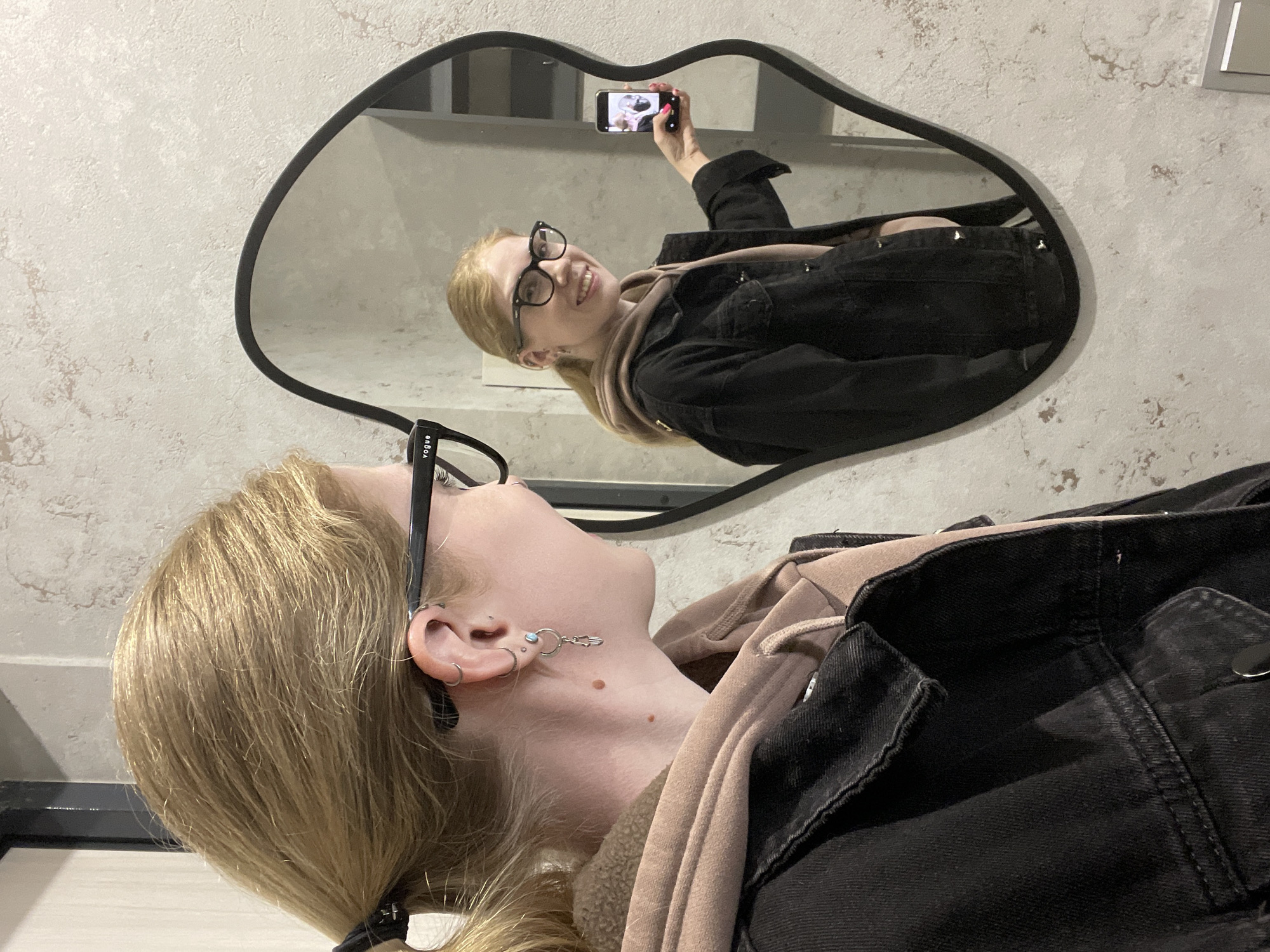 A young woman takes a selfie using her smartphone in a mirror with a distinctive lip-shaped frame. The mirror hangs on a wall with a speckled pattern, and the woman is wearing glasses, a beige hooded sweatshirt, and a black denim jacket. Her hair is blonde and pulled back into a ponytail. The reflection in the mirror shows the woman holding her phone, smiling, and taking a photo. No information available from the image to suggest it was taken in Izhevsk, Russia.