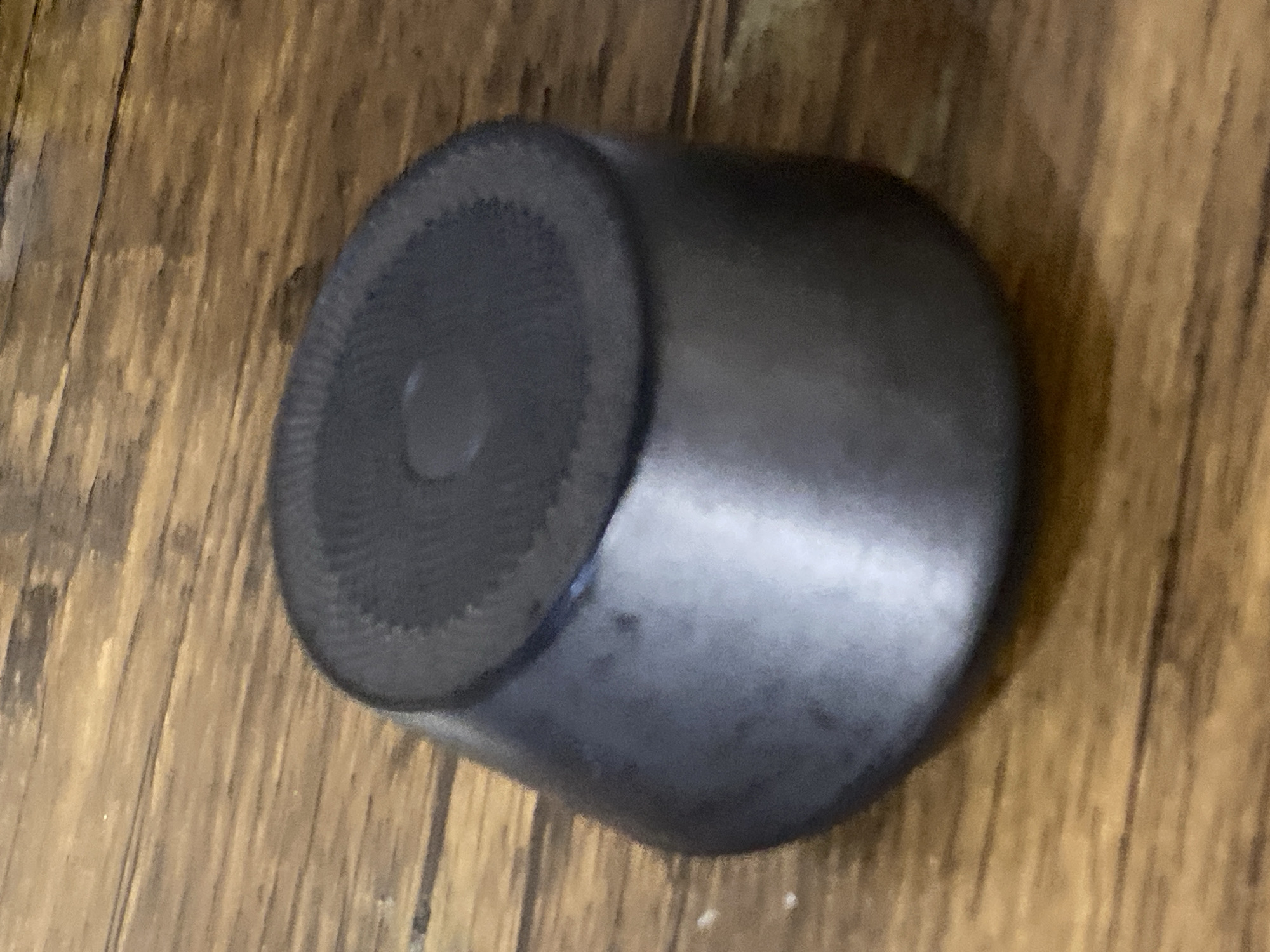 The image shows a dark, cylindrical object with a patterned top, resting on a wooden surface. The object appears to be made of metal and has a slightly tapered shape. The wooden surface is made up of planks with visible grain and some subtle variations in color. The lighting in the image suggests an indoor setting with a slight overhead light source, casting subtle shadows. There are no people visible in the image, nor are there any indications of specific actions or interactions. The image is a close-up shot of the object and its immediate surroundings. There is no information available to place this scene in Jauro Yinu, Nigeria.