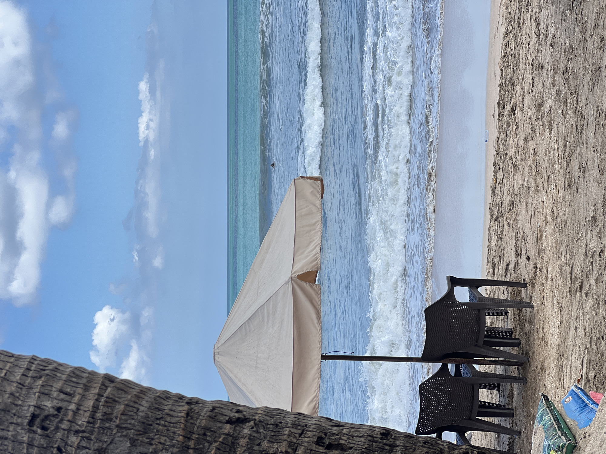The image depicts a tropical beach scene in Kuta, Indonesia, with the ocean stretching to the horizon under a partially cloudy sky. In the foreground, the rough texture of a palm tree trunk is visible on the left. Beneath a large, beige beach umbrella, two dark plastic chairs are positioned on the sandy shore. The ocean is active with moderate waves breaking, and a solitary surfer is visible in the distance. In the lower left corner of the frame, a green bag and a blue object are partially buried in the sand.