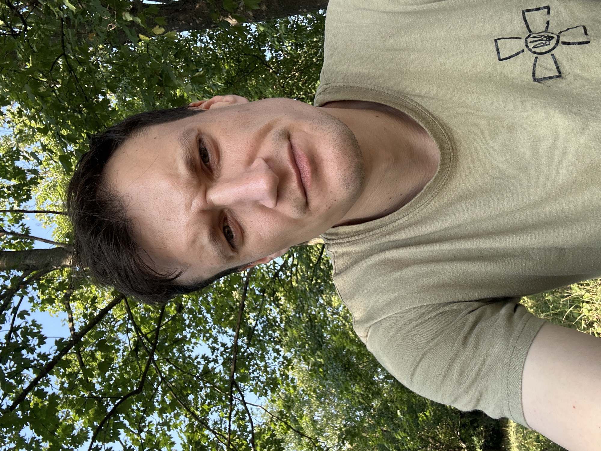 A close-up, selfie-style photograph shows a man with dark hair and a light complexion. He is looking directly at the camera with a slightly furrowed brow and a neutral expression. He is wearing a light olive-green t-shirt. On the left side of the t-shirt, a black emblem is visible, depicting a cross with a shield in the center. The man is outdoors, with lush green foliage and tree branches forming the background, suggesting a natural, possibly park-like setting. The lighting indicates it is daytime. There is no visible indication that the photograph was taken in Makiv, Ukraine.