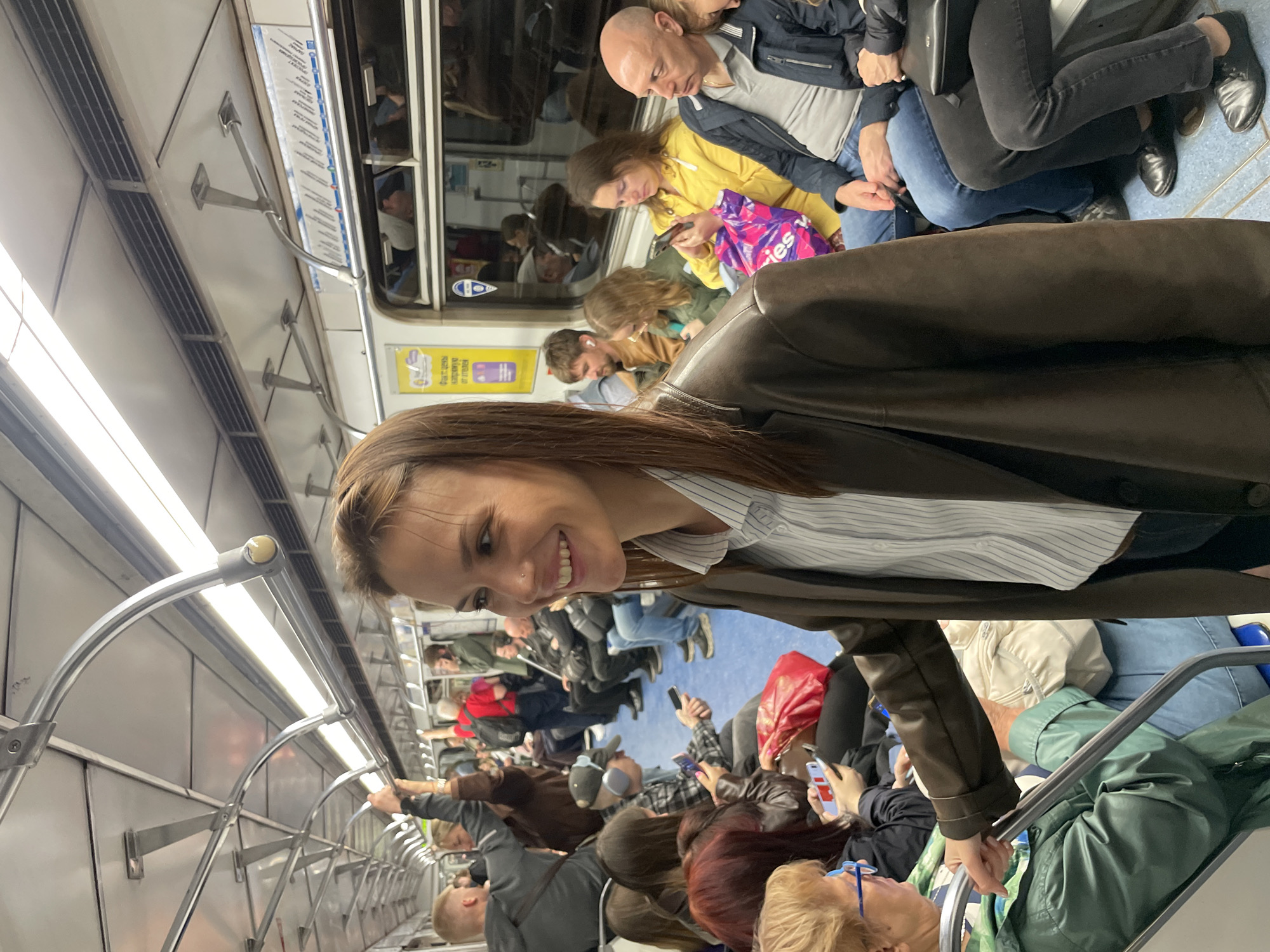 A crowded subway car is depicted, with numerous passengers either sitting or standing. Many passengers are holding or looking at their cell phones. Overhead, there are metal handrails running the length of the car, illuminated by fluorescent lights. A smiling woman wearing a brown blazer and a striped shirt looks directly at the camera. The city is not identifiable.