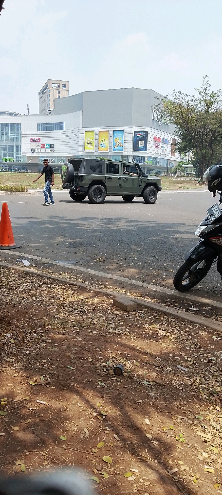 A man in a black shirt and blue jeans is standing on a paved road, gesturing with his right hand towards a dark green four-door vehicle with blacked-out windows, which is stopped on the road. The vehicle appears to be an off-road SUV with a spare tire mounted on the rear. In the background, a large, modern building with a gray facade and a large glass entrance is visible, identified by signs as AEON and various other store names. Trees and other vegetation are present in the background, suggesting an outdoor urban or suburban environment. An orange traffic cone is positioned on the left side of the road. The ground in the foreground is covered with dry leaves and dirt. Part of a black motorcycle is visible in the lower right corner of the image. The sky is clear and bright. The scene is set in Gunung Putri, Indonesia, likely near a commercial center.