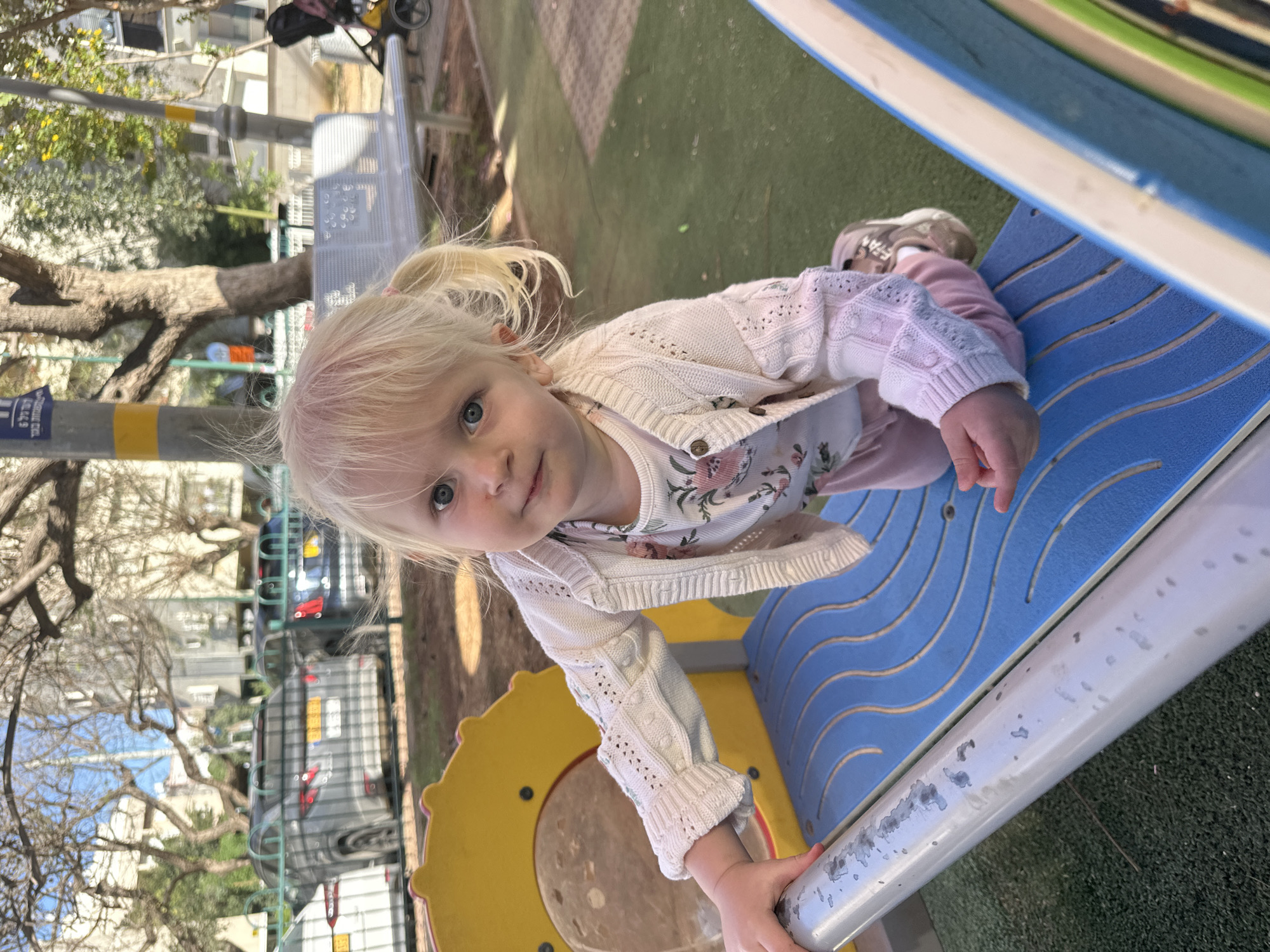 A young blonde-haired child with blue eyes looks directly at the camera while situated on a blue and yellow playground structure. The child, likely a girl, wears a light-colored floral top, a cream-colored knitted cardigan, and pink pants. Her blonde hair is styled in two small pigtails. She is holding onto a silver metal bar that is part of the playground equipment, which features wavy patterns on its blue surface. The ground surface of the playground is green artificial turf.
The setting is an outdoor urban or suburban environment, consistent with Petah Tikva, Israel, under bright daylight. In the background, beyond a fence, a street is visible with parked cars and multi-story buildings. Several trees, some bare and some with green leaves, line the street. A black stroller can also be seen in the distance. The child's expression is neutral and observant, as she engages with the viewer. Partial Hebrew text and numbers, including "בצרי", "ל", "4", and "9", are visible on a distant yellow and blue striped pole, serving as a location cue. The image is captured with a tilted perspective.