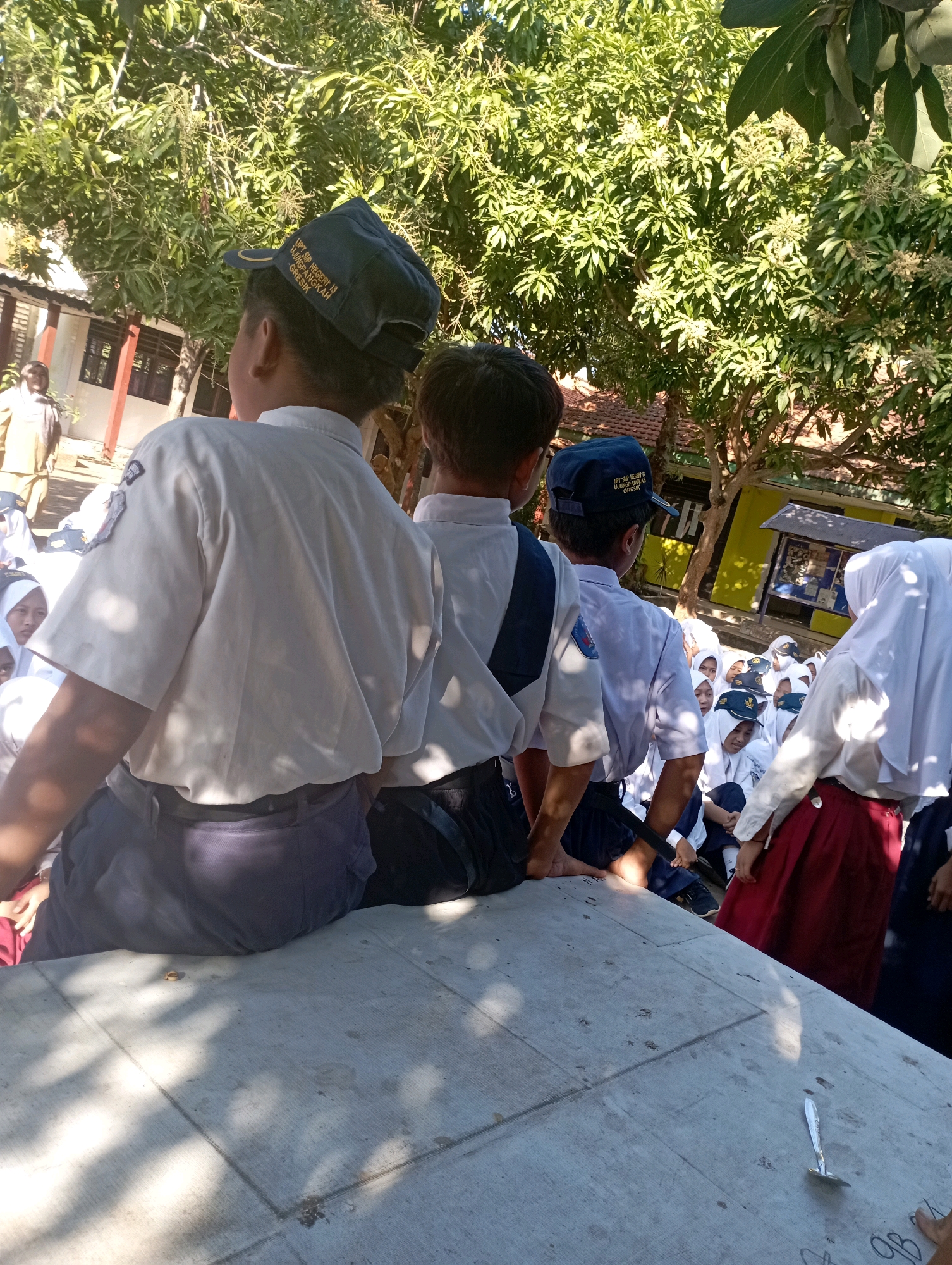 The image depicts a group of schoolchildren, predominantly wearing white shirts and dark trousers or skirts, in an outdoor setting. The scene appears to be within the grounds of a school, as indicated by the presence of buildings and trees. Several children are seated on a concrete surface, some facing away from the camera, while others are looking towards the left of the frame. One child in the foreground, facing away, is wearing a dark cap with yellow lettering that reads "UPT SDN NEGERI 18 Ujungpangkah Gresik." Another child, also facing away, wears a blue cap. A white building with red trim is visible in the background, along with mature trees providing shade. Some of the children are wearing hijabs. The overall atmosphere suggests a school activity or gathering. The location is identified as Banyuurip, Indonesia.