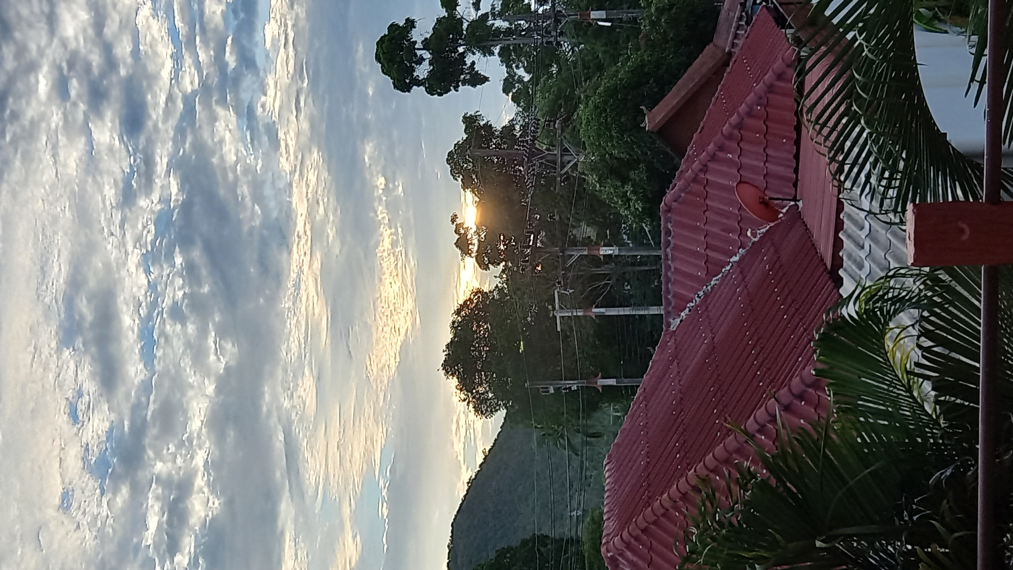 The image depicts a serene scene in the Ko Pha-ngan District, Thailand, during what appears to be either sunrise or sunset. In the foreground, the distinctive red-tiled roofs of several houses are visible, some adorned with satellite dishes. Lush green palm fronds frame the lower part of the image. Beyond the rooftops, a dense canopy of trees forms a verdant backdrop. In the distance, a rolling green hill is silhouetted against the sky. The sky itself is a canvas of dramatic clouds, with the sun’s rays piercing through, casting a warm glow. The overall impression is one of peaceful rural life in a tropical setting.