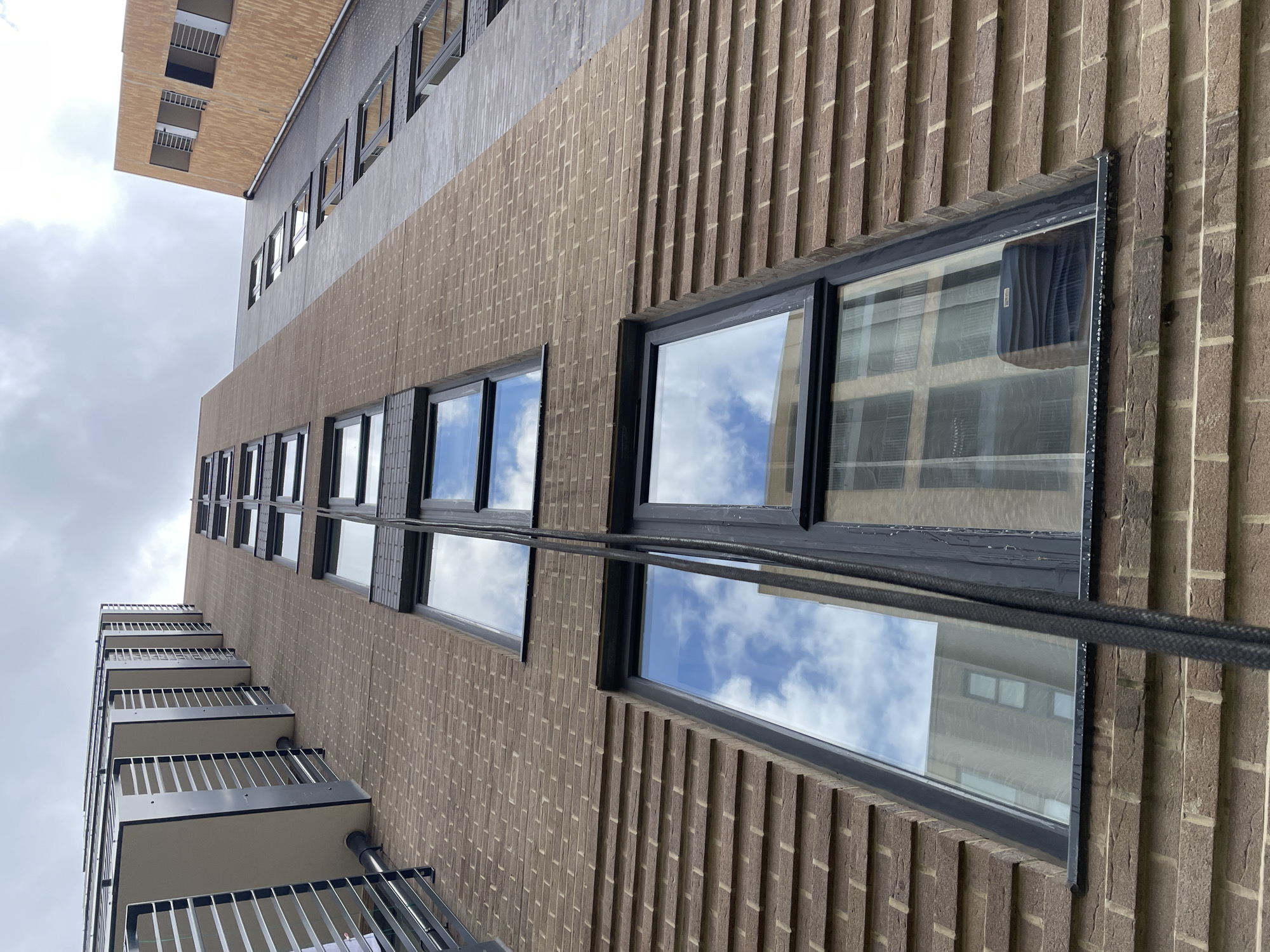 A low-angle shot focuses on a tall building's facade, potentially in London, UK, given the architecture. The building is predominantly brick, with a pattern of alternating vertical and horizontal bricks. A series of windows ascend the building's height, reflecting the sky and clouds. Some windows also reflect other buildings. A black cable runs vertically, close to the building's surface, intersecting with the windows. Balconies are visible on the building's left side, with dark metal railings. A section of the building at the top has a different brick color and texture. The sky is overcast with a mix of clouds and bright patches.