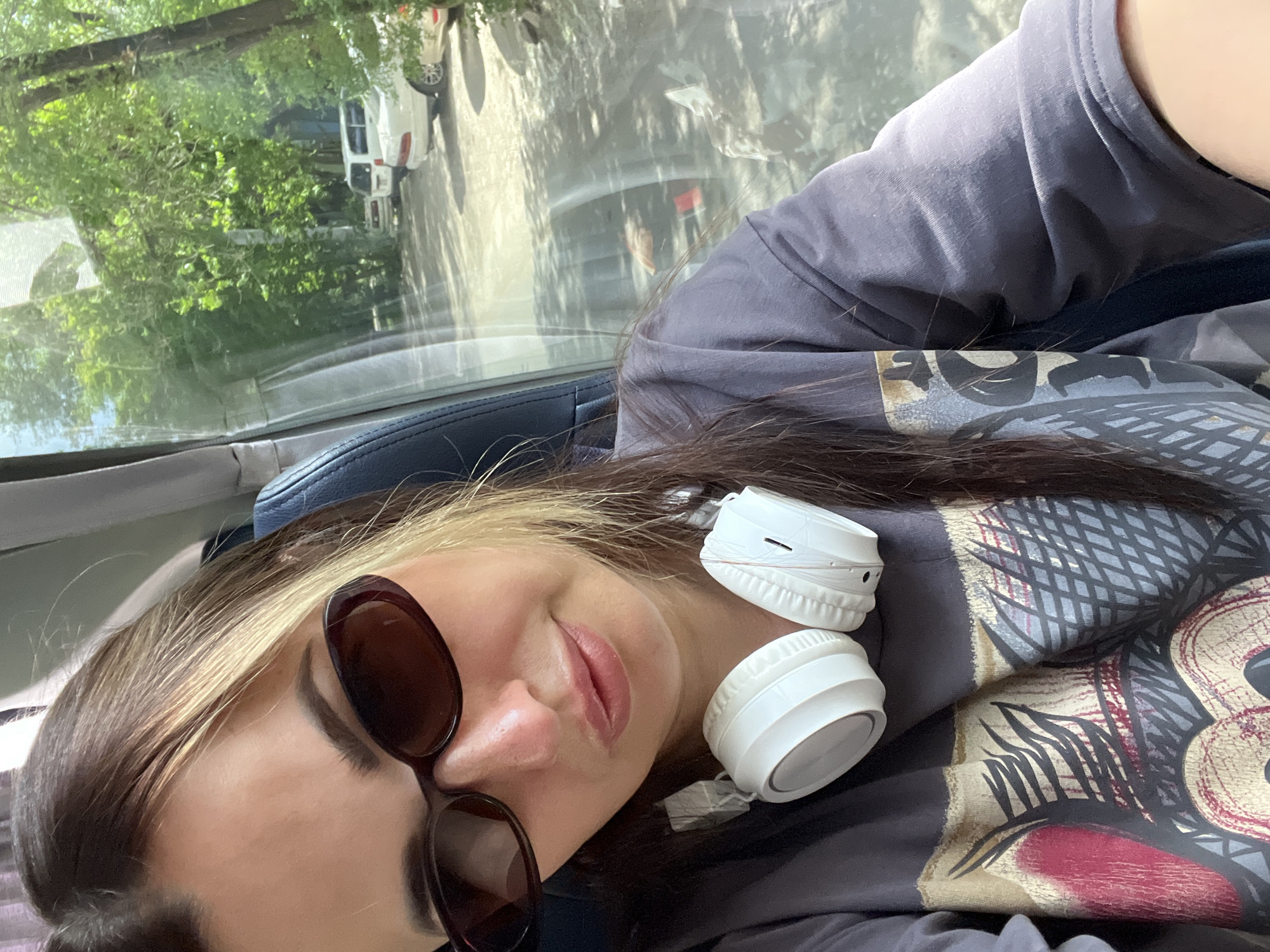 A medium shot shows a woman in sunglasses, wearing a dark gray t-shirt with a graphic print, and white headphones around her neck. She is seated inside a vehicle, with a window to her right offering a view of trees, parked cars, and dappled sunlight on the ground. The interior of the vehicle appears to be a bus or a van, with a dark blue seat visible behind her. The woman's hair is long and dark brown, with blonde highlights.