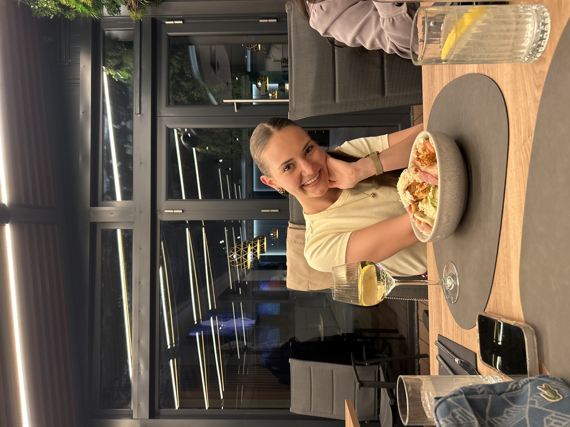 A young woman smiles at the camera while seated at a table in a restaurant. She is resting her face on her right hand. On the table is a bowl of salad, a glass of white wine with a lemon wedge, and a glass of water with a lemon slice. Silverware and a cell phone are also on the table. The restaurant features a wall of windows, offering a view of an illuminated building. The scene could be taking place in Sofia, Bulgaria.
