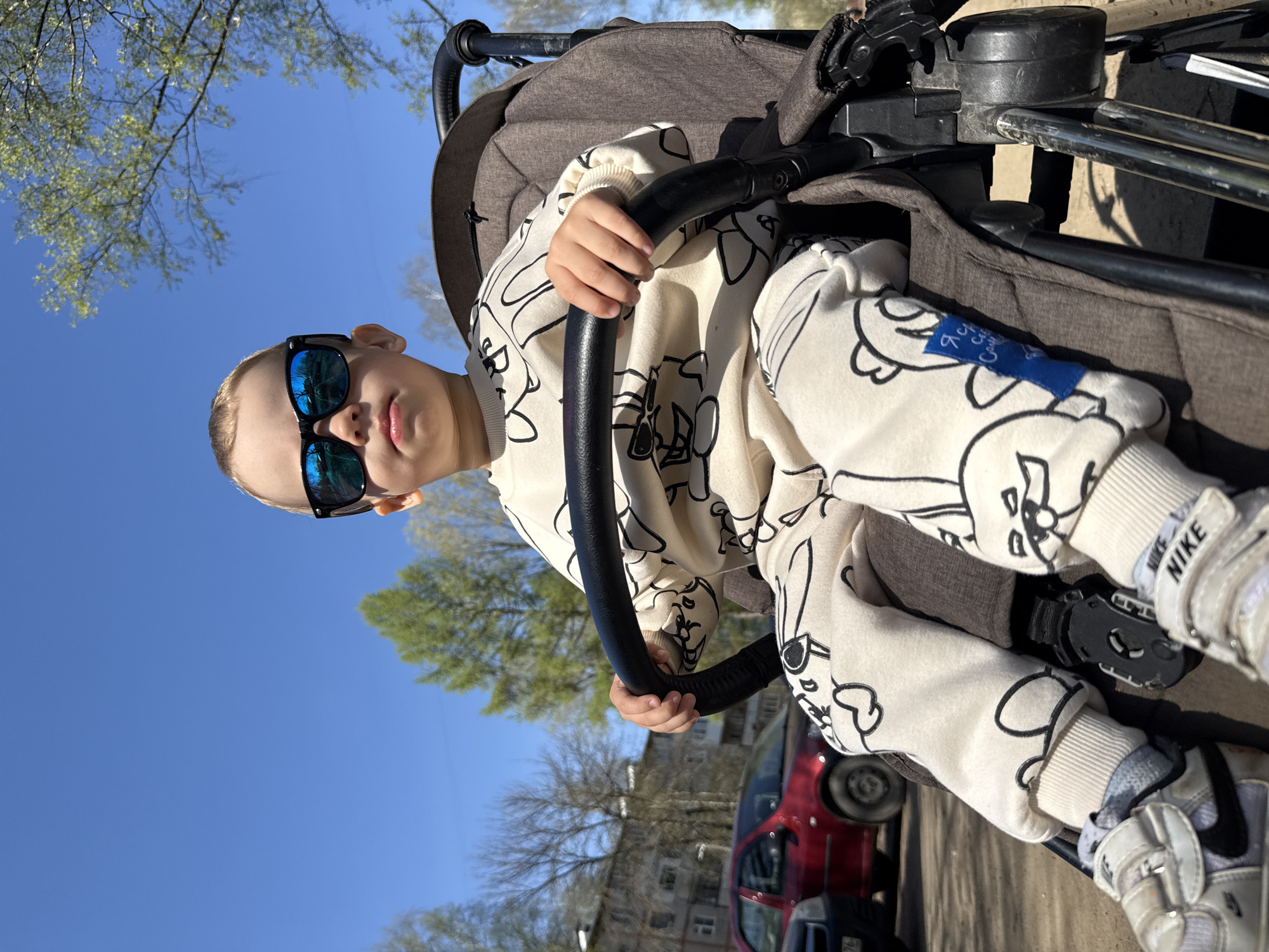 A low-angle, outdoor photograph captures a child in a stroller against a backdrop of blue sky and trees. The child, wearing dark sunglasses, is seated in a brown stroller, holding onto a black circular bar. The child is wearing a light-colored outfit patterned with dark cartoon figures. The child is also wearing white and gray Nike sneakers. Behind the stroller, a red car and a building are visible. The location is Rusanovo, Russia. The image is oriented vertically.
