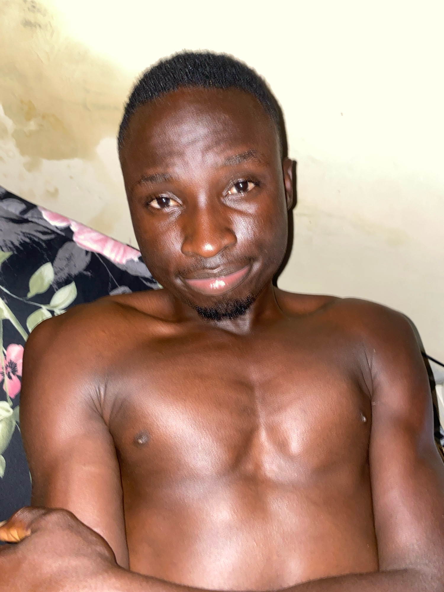 A young Black male with short dark hair and a light beard is shown from the chest up, lying down and looking towards the camera. He has dark brown skin and is shirtless. The background appears to be a wall with an off-white, textured surface and a floral patterned fabric to his left. The scene is lit from above and to the right, casting some shadows on his body. The image was taken indoors. There is no discernible information about the location in Barden Yana, Nigeria, within the image itself.
