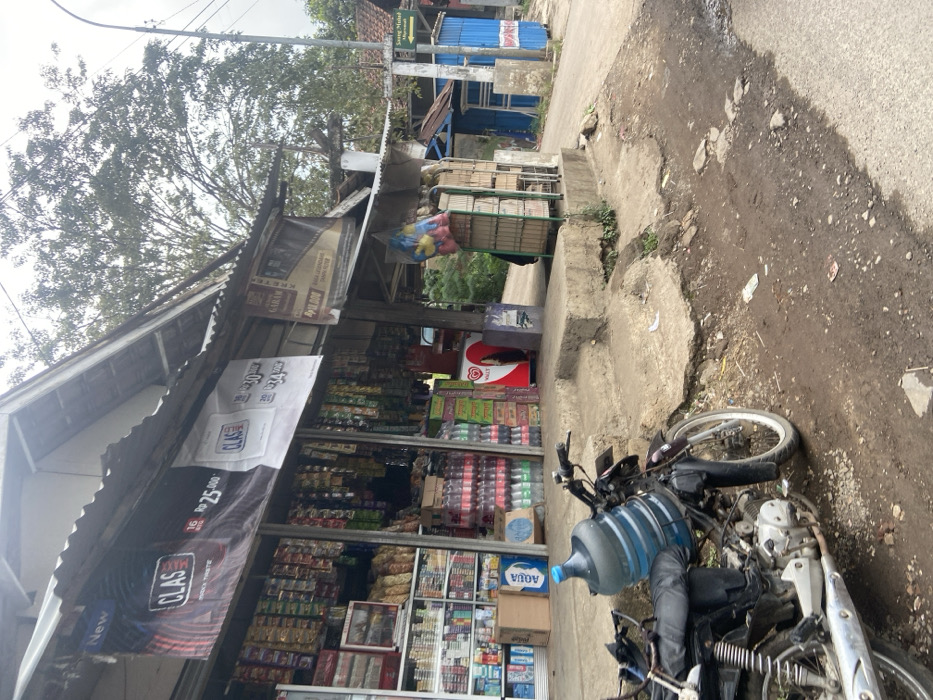 The image shows a small shop in Ciparay, Indonesia. The shop is located on the side of a dirt road and has a sign that says "CLAS" above the door. The shop is selling a variety of goods, including cigarettes, snacks, and drinks. A motorcycle is parked in front of the shop, with a large water bottle strapped to the back. There are two steps leading up to the shop door. The shop is surrounded by a small amount of greenery. A sign on the opposite side of the road reads, "Sungai Minda."