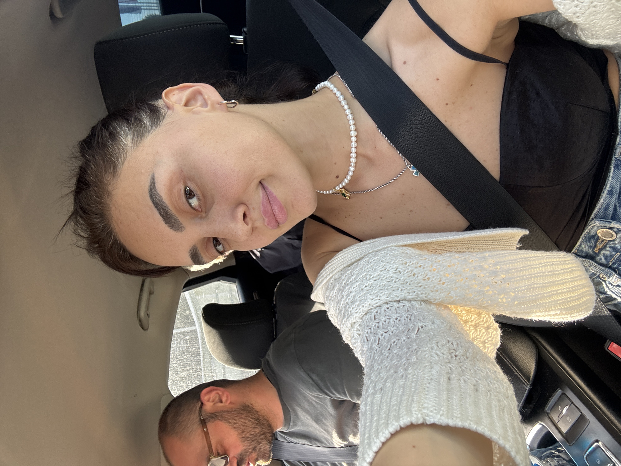 The image is an upside-down, interior shot taken inside a car. Two people are visible. The primary subject is a woman in the foreground, positioned on the right side of the frame. She has dark hair pulled back, prominent eyebrows, and is looking towards the camera with a slight smile. She is wearing a black top, a pearl necklace with a pendant, and a white knit cardigan. A black seatbelt crosses over her chest.
In the background, a man is visible on the left side of the frame, also appearing upside down. He has short hair, a beard, and is wearing glasses and a grey t-shirt. He is looking downwards.
The scene appears to be set during the daytime, with sunlight streaming into the car. The interior of the car is visible, including car seats, the dashboard, and a window showing an out-of-focus exterior. The location context is Tbilisi, Georgia. There is no visible text in the image. The overall mood seems casual and candid.