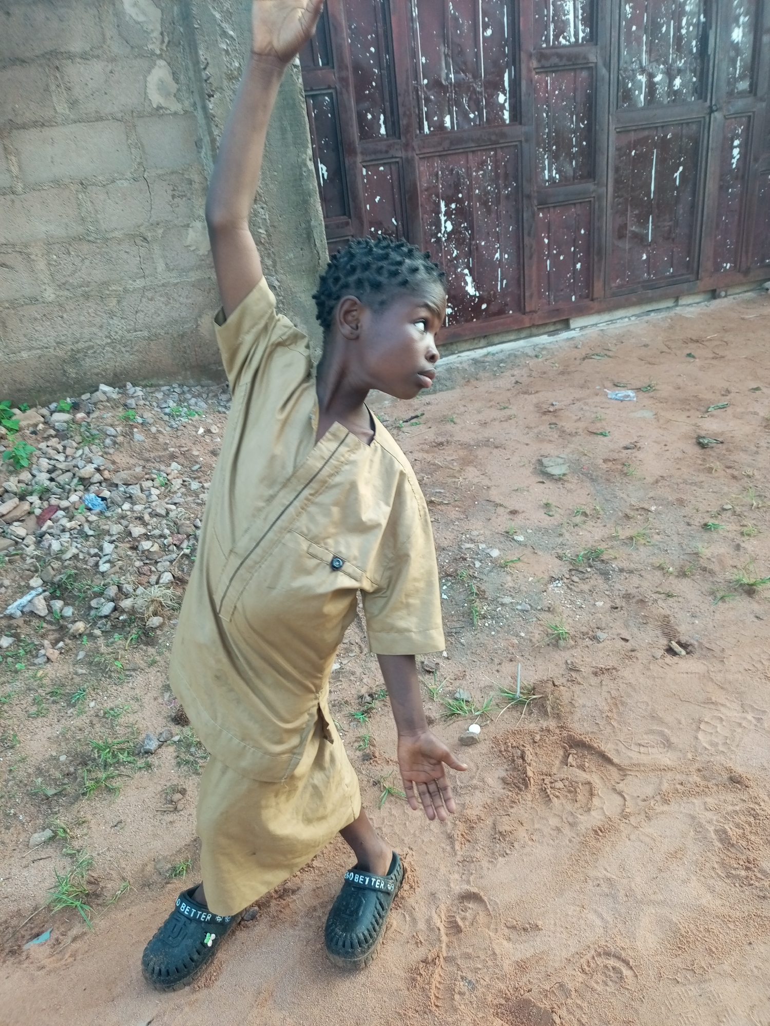 A young child, appearing to be of African descent, stands in a dirt and gravel area. The child is wearing a loose-fitting, light brown garment and black slip-on shoes with "BETTER" printed on them. The child's right arm is raised high above their head, and their left hand is extended palm up. The child's head is turned to the right, and they appear to be looking towards something out of frame. In the background, there is a textured brick wall and a dark, weathered metal door or gate. The ground is sandy with scattered stones and sparse green vegetation. The scene is outdoors, with natural lighting suggesting it is daytime. The location is identified as Tempure, Nigeria.